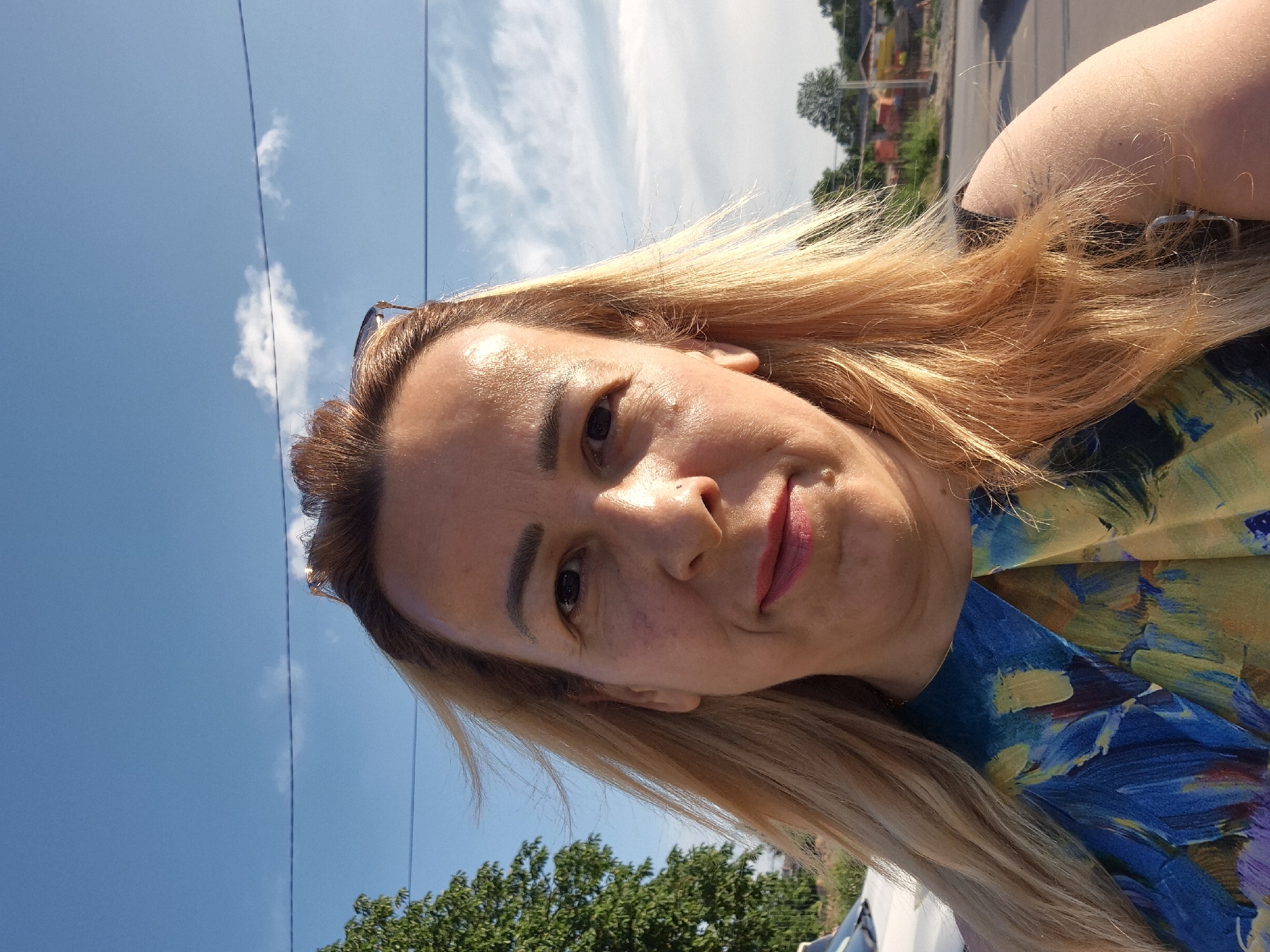 A woman is taking a selfie outdoors on a sunny day. She has blonde hair and is wearing a blue and yellow patterned shirt. The background shows a blue sky with some clouds, trees, and a road.