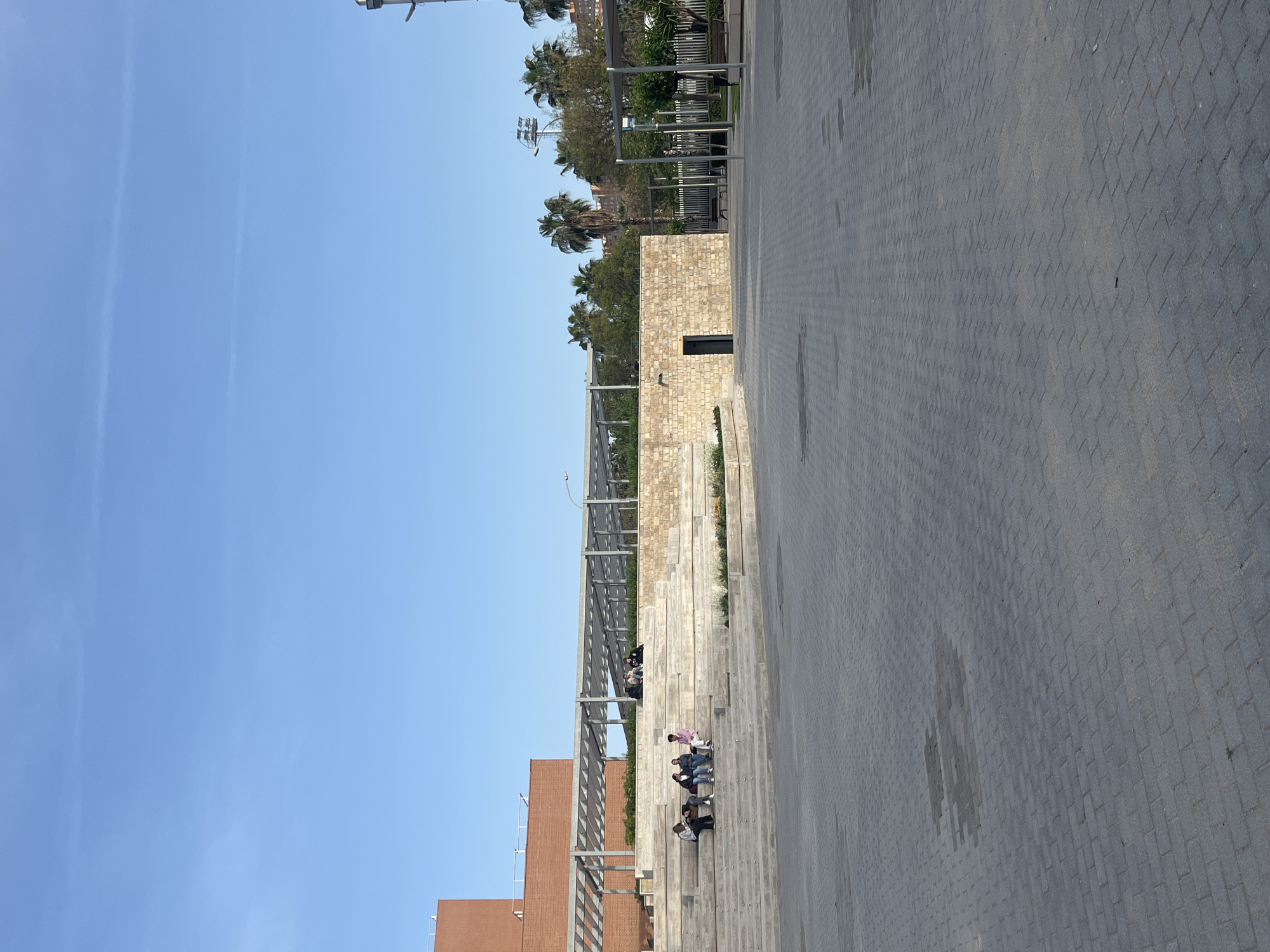 This image depicts a modern urban public space in València, Spain, under a bright, clear blue sky with faint contrails. The foreground and right side are dominated by a wide plaza paved with grey interlocking paver blocks, creating a textured ground surface.
To the left, a series of tiered concrete or stone steps forms an outdoor seating area or amphitheater. Several individuals are visible sitting casually on these steps, scattered across different levels. They appear to be relaxing or socializing, dressed in everyday attire, with one person prominent in a light pink top.
Above the seating tiers, a long, open metal pergola or trellis structure spans horizontally, suggesting a shaded walkway or resting spot. Behind this structure and the steps, a landscaped area features lush greenery, including multiple palm trees, indicating a warm climate. In the mid-ground, a modern building constructed of light-colored stone or brick stands, characterized by a dark, rectangular opening. A portion of another reddish-brown building is visible further to the left, and a tall light pole with fixtures extends into the sky on the upper right.
The overall scene suggests a pleasant, sunny day in a contemporary public square designed for leisure and community gathering. There is no visible text in the image.