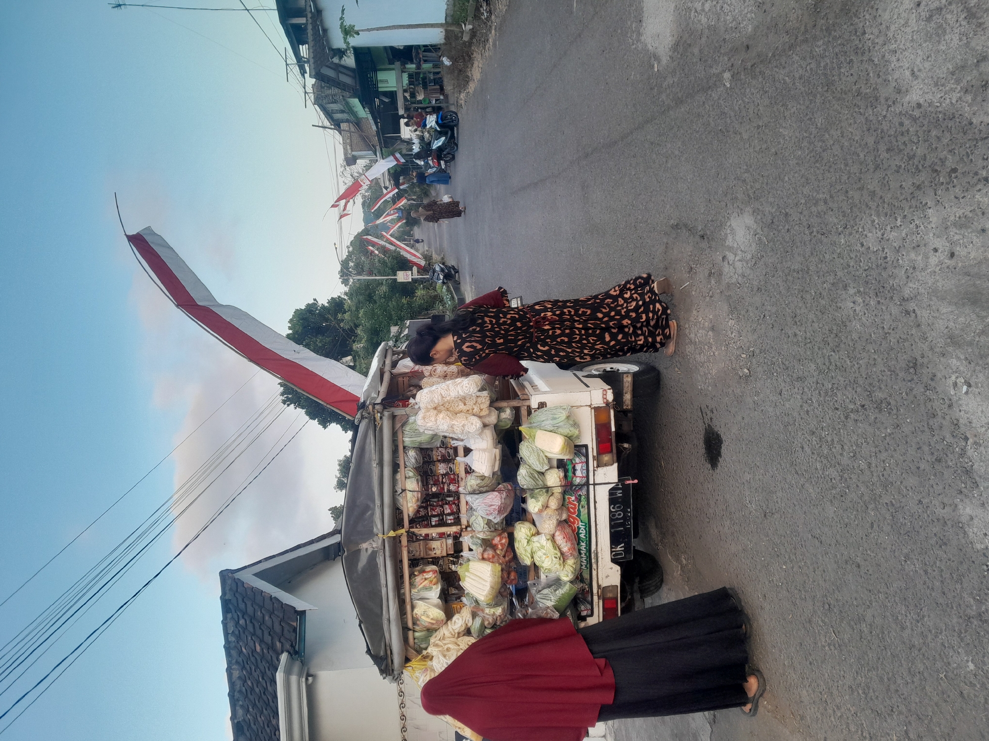 The image depicts a street scene in Sempu, Indonesia, under a pale blue sky. A white pickup truck, laden with produce and packaged goods, is parked on the side of the road. The truck's bed is filled with various items, including leafy vegetables in plastic bags, bundled produce, and numerous packaged snacks. A woman wearing a dark patterned dress stands beside the truck, facing away from the camera, appearing to interact with the merchandise. Another individual, draped in a dark red hijab and black attire, stands to the left of the truck, also facing away.
Along the street, red and white Indonesian flags are prominently displayed, stretched across the road at intervals, suggesting a celebratory or national event. In the background, more flags are visible, as are several buildings lining the street, some appearing to be shops or residences. A few motorcycles are parked on the side of the road, and other individuals can be seen walking in the distance. The road surface is asphalt, with some patches of lighter grey. The overall impression is a bustling street scene in a semi-urban or rural Indonesian setting, possibly during a national holiday.