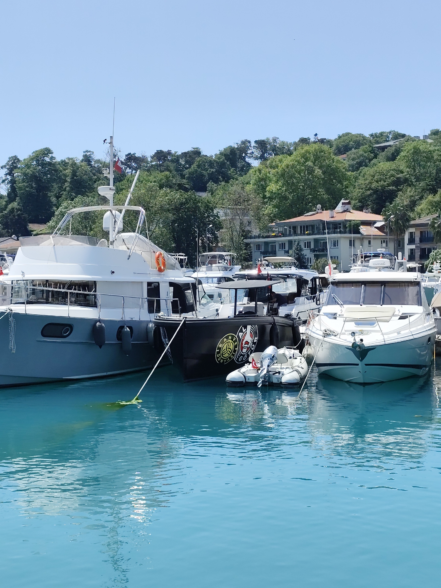 This vibrant photograph captures a serene marina scene in Sarıyer, Turkey, under a brilliant, clear blue sky. The image focuses on several boats docked in exceptionally clear, calm turquoise waters.
In the foreground, a large, modern grey and white yacht is prominently featured on the left, adorned with fenders and an orange life preserver. Its mast includes various antennas and a small red and white flag. Text, likely "Raymarine," is visible on equipment mounted on this yacht. To its right, a sleek black-hulled boat is moored, displaying a distinctive circular graphic with text that reads "KARAHAN DÜNYASI GÖL". Nestled between the black boat and the white yacht on the right is a small inflatable dinghy equipped with an outboard motor. The white yacht, with its clean design, also features navigational gear, with the brand "GARMIN" clearly visible on its dashboard.
The background showcases a verdant hillside densely covered with lush green trees. Several residential buildings with terracotta roofs and balconies are nestled within the greenery, overlooking the water. The bright sunlight and lack of harsh shadows suggest it is a clear, sunny mid-day. The water beautifully reflects the hulls of the boats. No people are visible in the frame, allowing the focus to remain on the boats and the picturesque natural setting. The overall atmosphere conveys a sense of calm and leisure in a beautiful coastal environment.