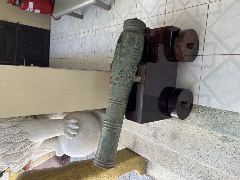 An old cannon rests on a dark wooden carriage with circular wheels. The cannon itself is green, possibly bronze, with ornate designs. It sits beside a marble statue of a figure holding a globe. Behind the cannon, a tan wall with a white-tiled floor is visible. A red fire extinguisher is mounted to the left of the scene, above a black door. The scene is brightly lit.