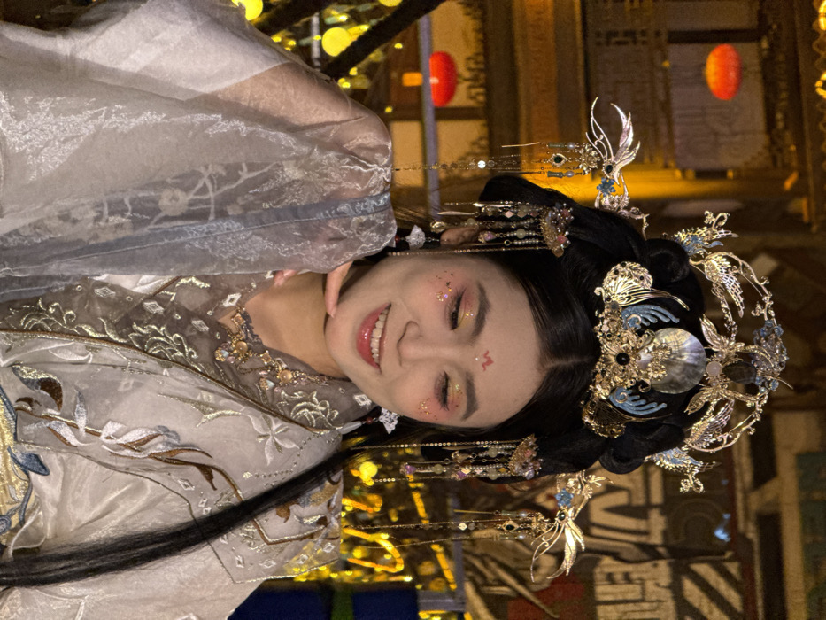 A medium-shot portrait depicts an East Asian woman, situated in Yuzhong District, China, adorned in elaborate traditional Chinese attire. Her dark hair is styled in an ornate updo, surmounted by a complex golden headpiece embellished with jewels, pearls, and metallic bird motifs, featuring multiple dangling ornaments. Complementary long, beaded decorative elements hang from her hair on both sides of her face. Her makeup includes prominent pink and gold eyeshadow with glitter accents on her upper cheeks, a small red mark centrally placed on her forehead, and glossy pink lips forming a gentle smile that reveals her teeth. She wears layered traditional garments in white and grayish-blue hues, including a sheer outer robe with subtle embroidery and an inner garment detailed with metallic, possibly gold, patterns around the neckline. Her right hand is raised, with her chin resting on her fingertips in a graceful pose. The background is softly blurred, showcasing warm, golden artificial lighting, traditional Chinese architectural elements, and two clearly visible, illuminated red lanterns, suggesting an evening or nighttime environment.