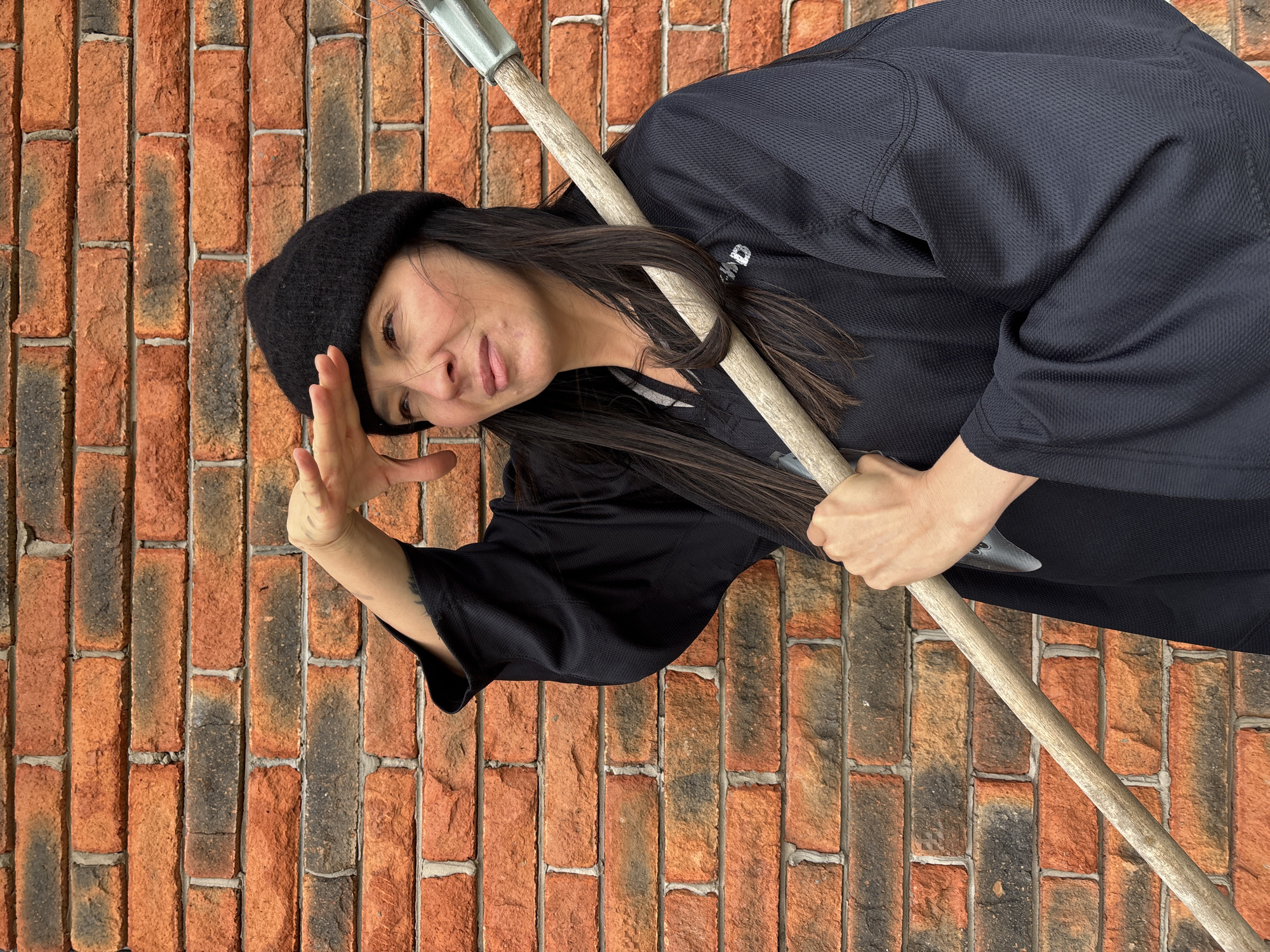 An adult woman with long dark hair and a black beanie is depicted in a medium shot against a textured brick wall. She wears a dark, possibly black, long-sleeved top or jacket made of a textured fabric, with partially visible white text, "AD", on the sleeve. She holds a long, light-colored wooden staff with a metallic tip across her body. Her left hand is raised to her forehead, and she displays a slight smile, conveying a playful or observant expression. The image is rotated 90 degrees clockwise, presenting the woman horizontally as if leaning against the wall or caught in motion. The background features a brick wall composed of varied shades of reddish-orange and dark grey bricks with light mortar lines. The lighting suggests a bright, possibly overcast, daytime setting. The scene is set in Chaikovsky, Russia.