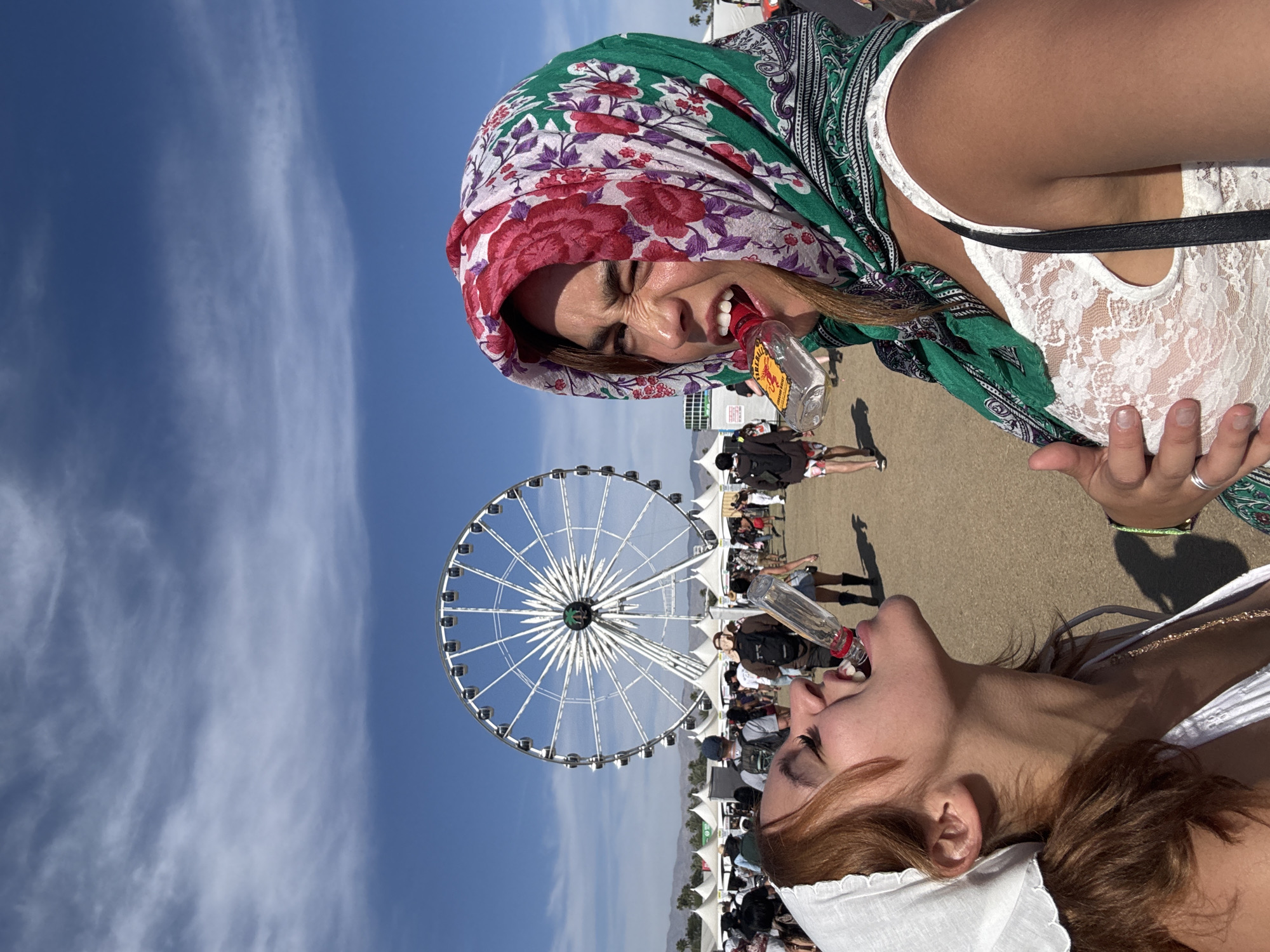 Two young women are captured in a vertical, close-up shot at an outdoor festival. The top woman, wearing a vibrant green, red, and purple floral headscarf over brown hair and a white lace top, holds a small clear bottle to her mouth. Her face is contorted in an exaggerated expression, with eyes tightly scrunched and mouth wide open, suggesting a strong reaction to the drink. A red and yellow label on her bottle is partially visible, appearing to be from the Fireball cinnamon whisky brand. Below her, partially visible, another woman with reddish-brown hair, wearing a white top and a white head covering (possibly a bandana), also drinks from a similar small clear bottle, tilting her head back with her eyes closed.

In the background, under a bright blue sky with wispy clouds, a large white Ferris wheel stands prominently. The scene unfolds on what appears to be dusty ground, with numerous people and white tents scattered across a festival site. The sunny daytime weather and visible festival elements, coupled with the location context of Indio, USA, strongly suggest the Coachella Valley Music and Arts Festival. The women's animated expressions convey a sense of fun and excitement, indicative of the festive atmosphere.