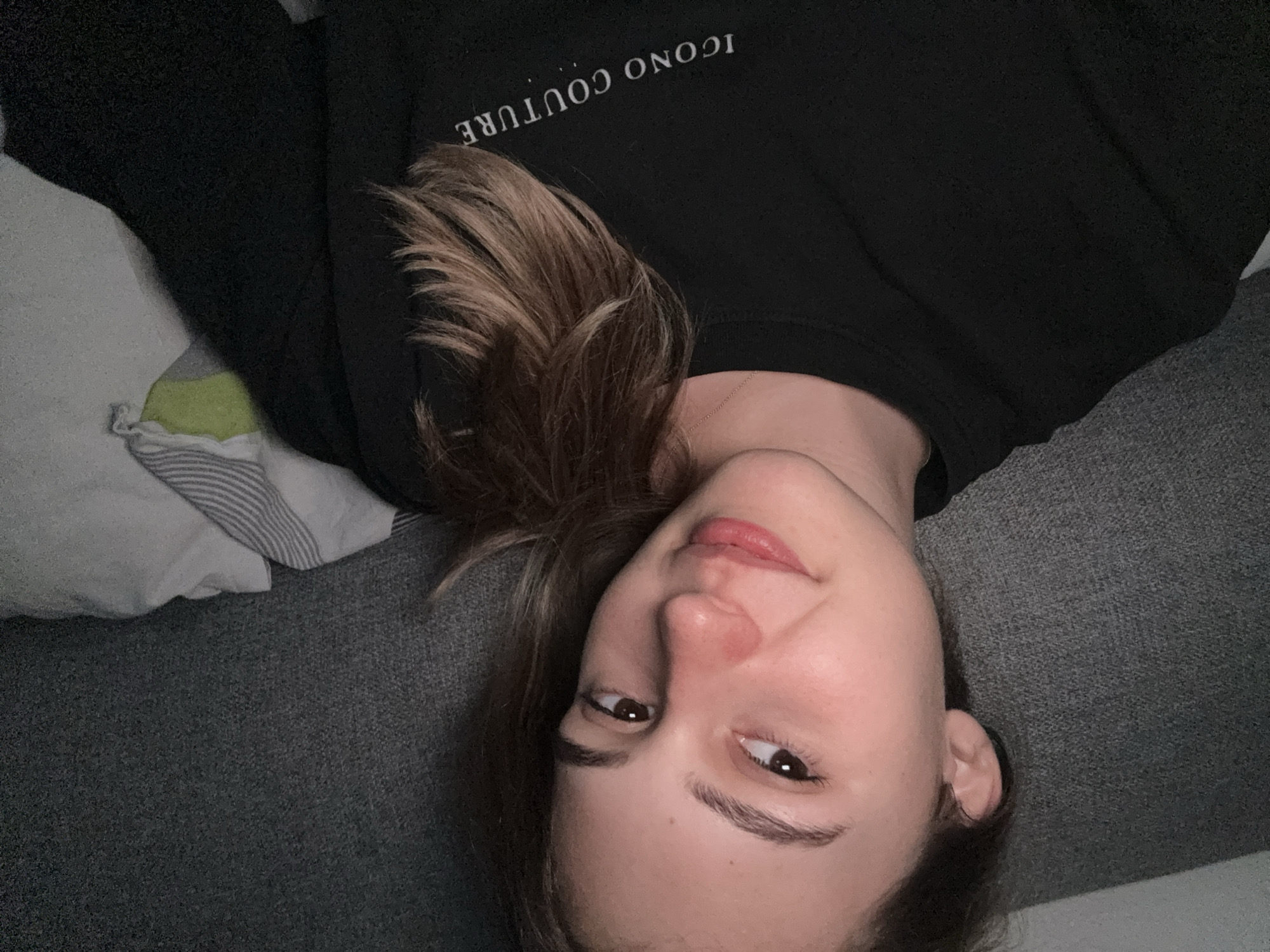 A young woman with light brown hair and fair skin is shown in a close-up selfie. She is wearing a black sweatshirt with the text "ICONO COUTURE" printed in white on the chest. The woman is lying down, leaning against a textured grey surface, possibly a couch or bed. Her hair is partially covering her right shoulder. To her right, a white pillow with grey stripes and a lime green corner is visible. The lighting appears to be artificial and subdued. The scene does not provide any identifiable landmarks or features that would indicate a specific location within the city of Forbach, Germany.