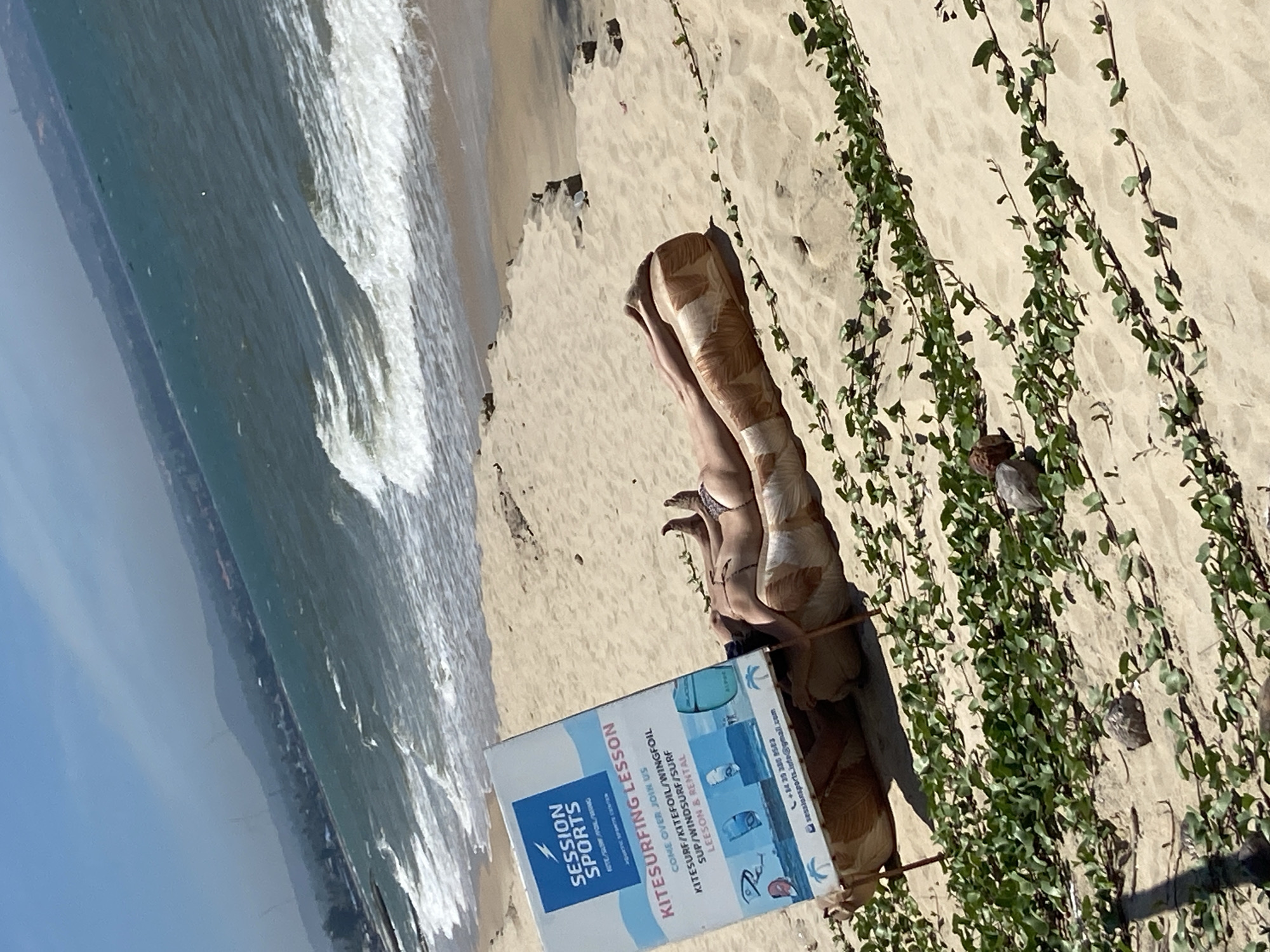 This media file depicts a bright, sunny day at a beach in Phu Thuy Ward, Vietnam. The scene features a sandy beach with the ocean and waves on the left side. Green vines with numerous leaves are growing along the sand dunes, casting shadows. In the foreground, a blue and white sign advertises "SESSION SPORTS" and "KITESURFING LESSON," listing activities like kitesurf, kitefoil, wingfoil, SUP, windsurf, and surf, along with lesson and rental services. Contact information, including a phone number and email address, is visible on the sign. Partially obscured by the sign and lying on the sand are the legs of a person wearing a swimsuit and a patterned wrap. The person appears to be relaxing or sunbathing. The image is taken at an angle, with the shore and waves running diagonally across the frame. The overall impression is a relaxed beach environment offering water sports.