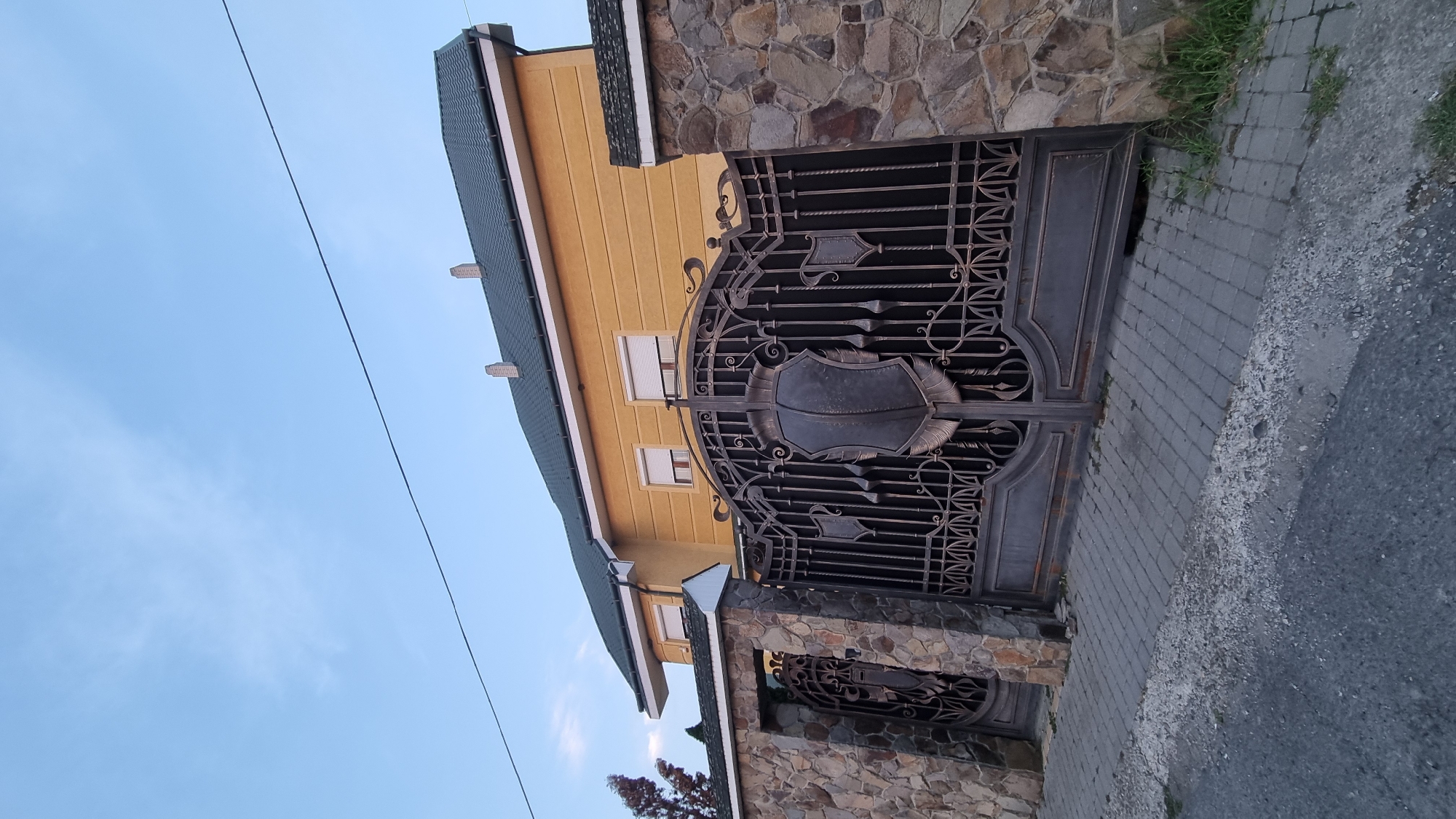 An exterior, low-angle, eye-level shot captures a large, ornate wrought-iron gate in front of a yellow, two-story house. The gate, dark gray and intricately designed with decorative elements including shield-like motifs, is flanked by a stone wall on the left and a similar wall on the right, partially obscured by the gate. To the left of the main gate, a smaller, similarly designed wrought-iron pedestrian gate is visible within the stone wall. The house features a green, tiled roof with two brick chimneys. Its yellow facade has several windows covered by white blinds or shutters. The ground in front of the gate is paved with gray interlocking bricks, transitioning to asphalt on the left side. The sky above is a clear, pale blue with sparse, wispy clouds. The location is identified as Berehove, Ukraine.