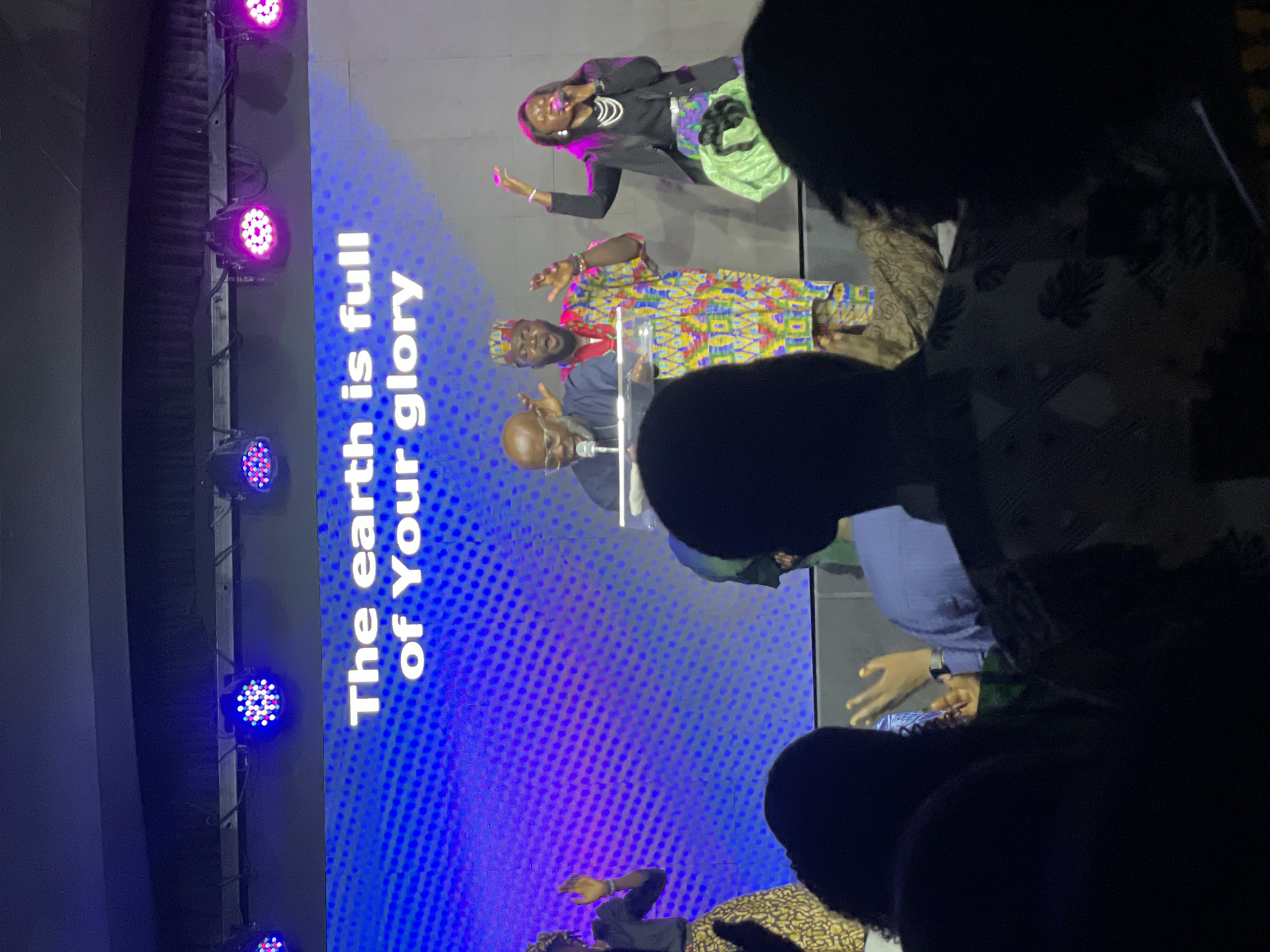 A photo taken in Jos, Nigeria, shows a stage setup with three speakers standing behind a lectern in front of a large screen. The screen displays the text "The earth is full of Your glory" against a blue and purple dotted background. The speakers include an older man with glasses standing at the lectern, a younger man in a colorful shirt, and a woman in a black blazer. Audience members are visible in the foreground. Stage lighting fixtures line the left side of the screen.