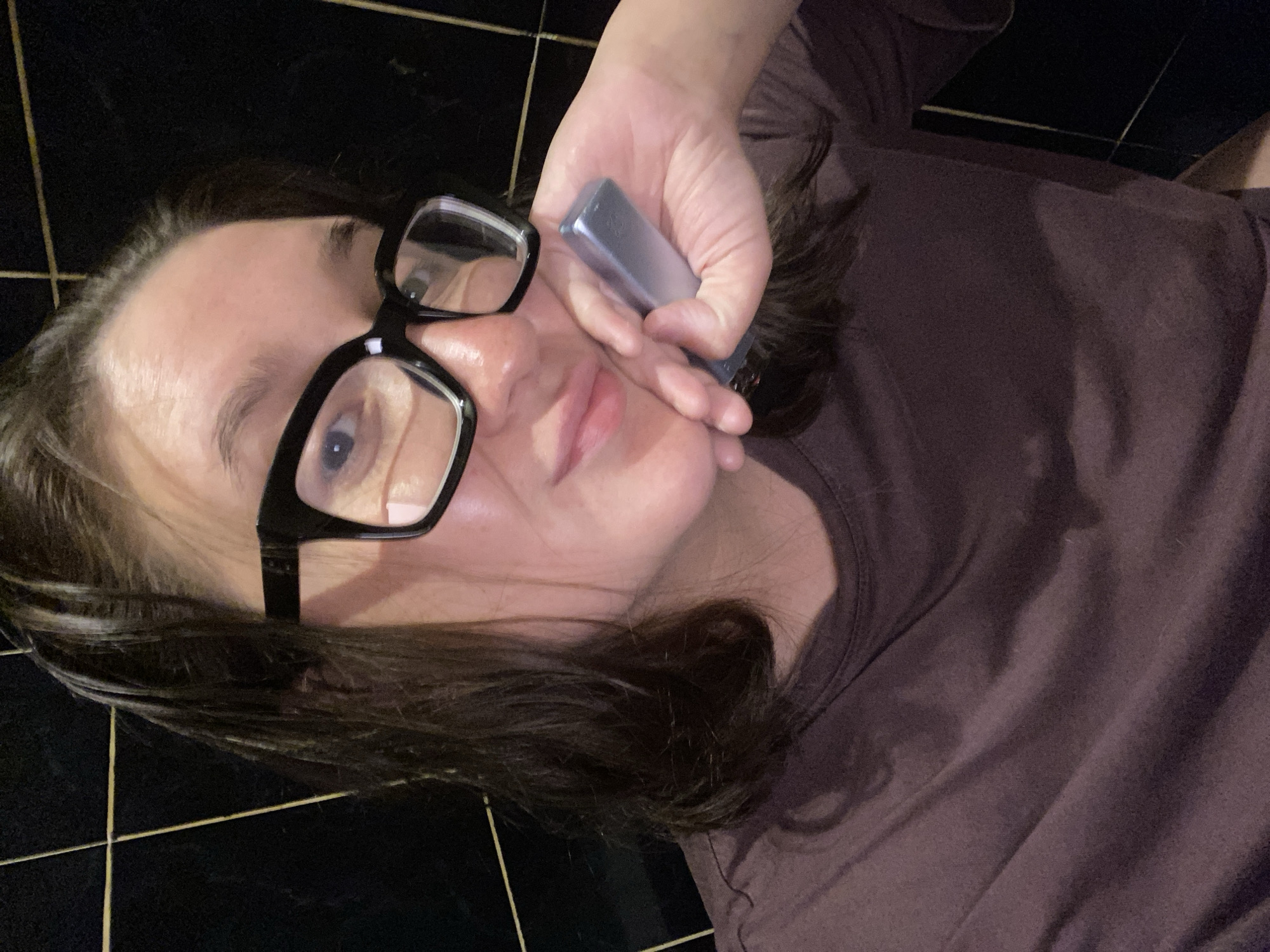 A young woman with dark hair pulled back is looking at the camera, wearing large black-framed glasses and a maroon t-shirt. She is resting her head on her hand, which is holding a small, metallic object. The background appears to be a dark, tiled wall. The image does not provide any information about a specific location in the city of Chaikovsky, Russia.