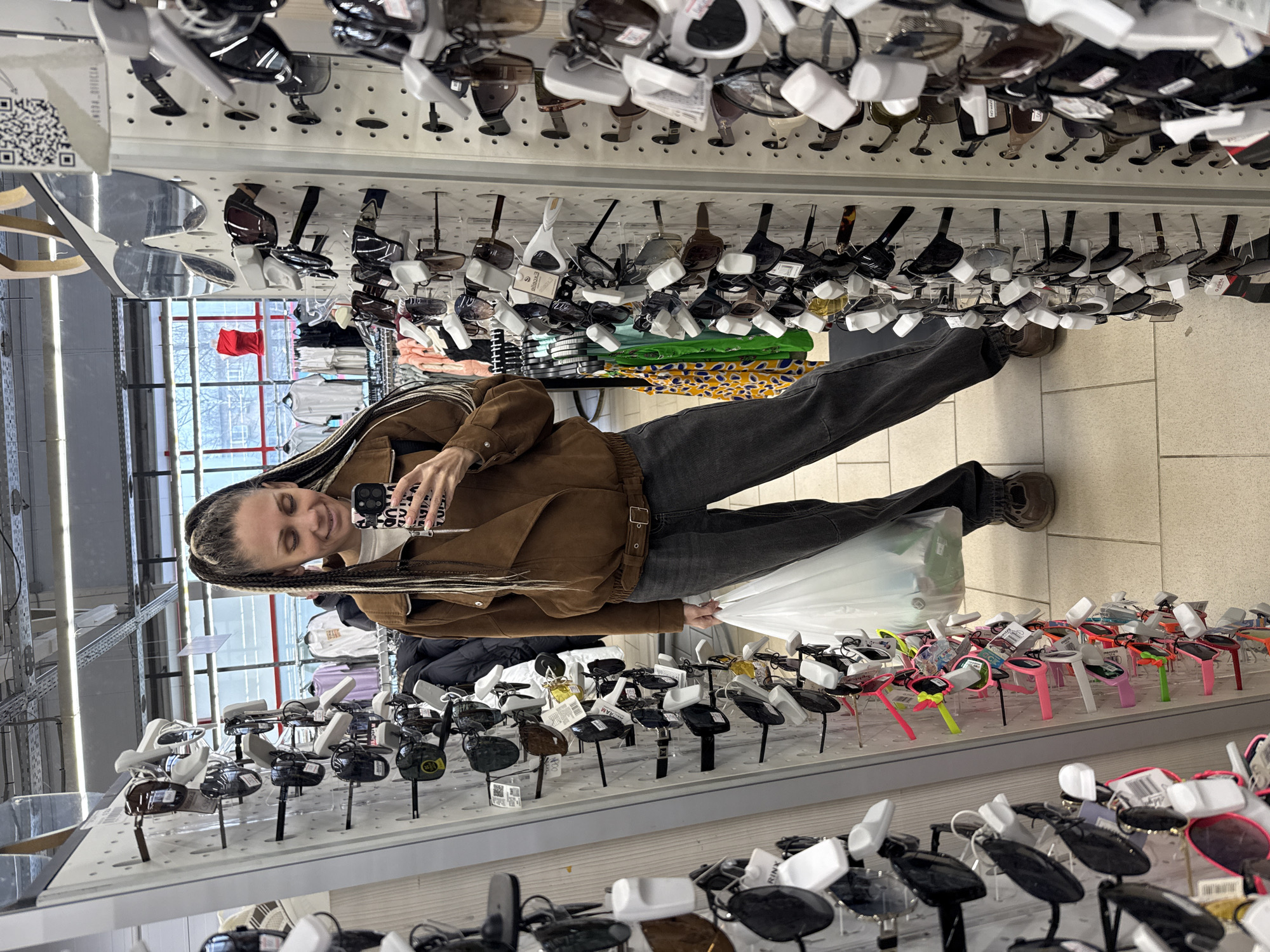 A woman is taking a selfie with her phone in what appears to be a store. She is standing in front of displays of sunglasses and possibly other items. The location is possibly Rusanovo, Russia. She is wearing a brown jacket and dark jeans and holding a white plastic bag. The store has bright lighting and a wide variety of merchandise on display.