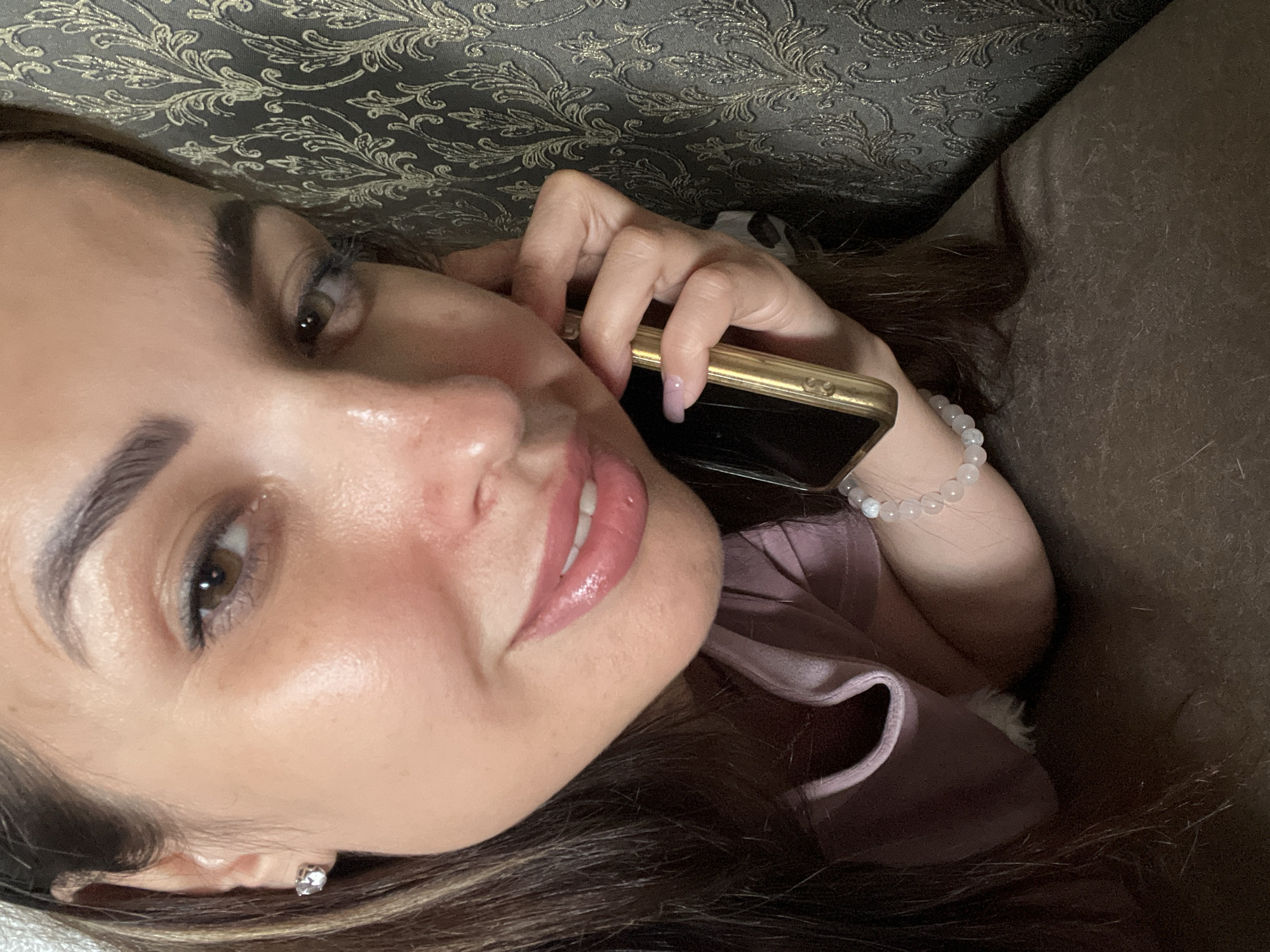 In a close-up, indoor shot, a woman is shown holding a gold-colored smartphone to her right ear and speaking. She is lying on her side on a brown surface, with her head tilted slightly upwards. Her hair, which is dark brown and long, is spread out around her. She is wearing a pale pink top and a bracelet made of white beads on her left wrist. Her right hand is holding the phone, and her nails are painted a pale pink. The background consists of a dark gray textured wall with a gold floral pattern. The lighting in the image is soft and appears to be coming from the front. The woman's face is turned towards the camera, and she is smiling slightly.