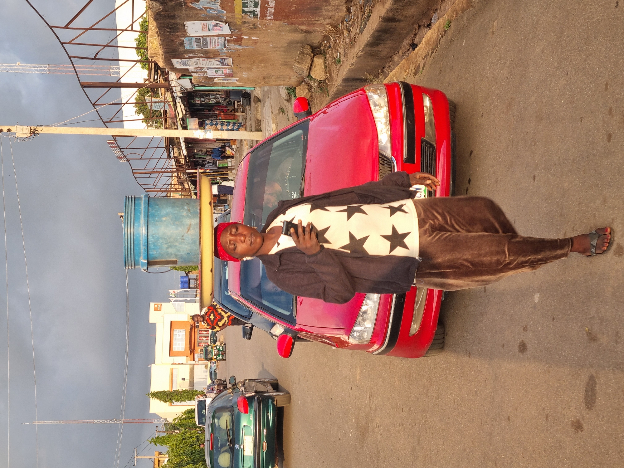 A man stands next to a red car on a street in Jos, Nigeria. The man wears a white shirt with black stars, a black jacket, and brown pants. He holds a mobile phone in his hand. The street is paved and appears to be a commercial area with shops and other cars visible in the background. The sky is overcast. A blue water tank is also visible.