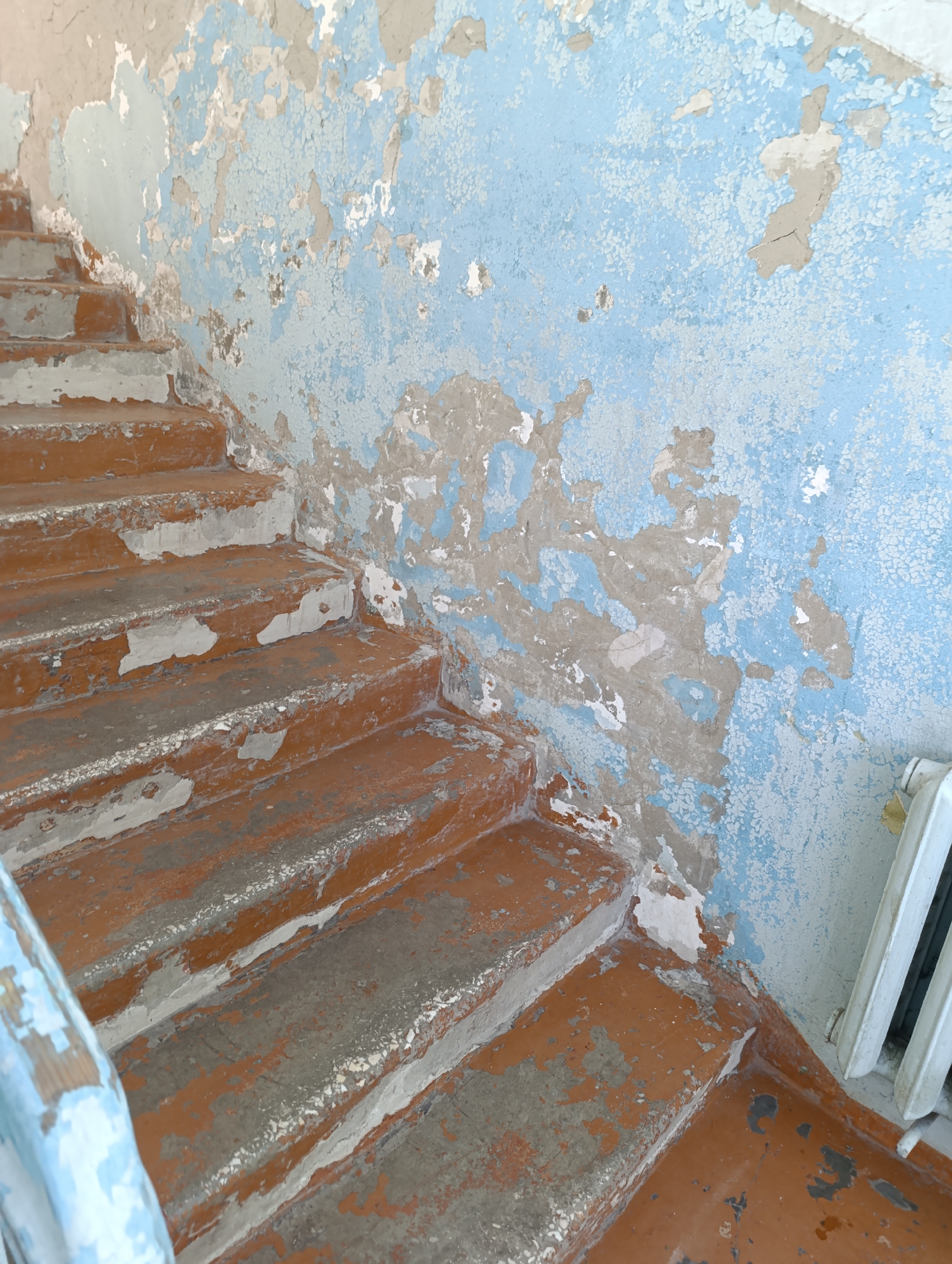 The image depicts a section of an interior staircase, likely within an apartment building or a public institution, characterized by visible signs of wear and disrepair. The staircase itself features concrete steps with worn, reddish-brown risers and treads. The surfaces of the steps are chipped and peeling, revealing the underlying concrete. A light blue handrail, also showing signs of peeling paint, is partially visible on the left side of the frame.
The wall adjacent to the staircase is painted a light blue color, but much of it has deteriorated, with large sections of paint flaking off, exposing a beige or grey underlayer. This peeling paint creates a textured and uneven surface on the wall.
In the background, on the right side of the image, a white radiator is visible, suggesting the presence of a heating system. The overall impression is one of an aged and neglected interior space. The location is identified as Tugulym, Russia, though no specific context is provided to pinpoint the exact building or its function within the town. There are no people, animals, or any indications of recent activity visible in the image.