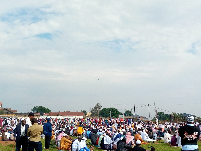 The image shows a large gathering of people, possibly a religious assembly or a public event, taking place on a grassy field in Beni, Democratic Republic of Congo. The location is likely a public space, possibly a park or a field adjacent to a mosque, as indicated by the presence of the people and the general setting. There are over 100 people present, mostly men, wearing various clothing, some with traditional white headwear, others with casual attire, and a few with a hoodie and jeans. The attendees are seated on the ground, engaging in conversation and prayer. The field is mostly covered with green grass with a few visible trees in the background, indicating an outdoor setting. The sky is cloudy with some sunlight. It appears to be a sunny day in the afternoon or evening.