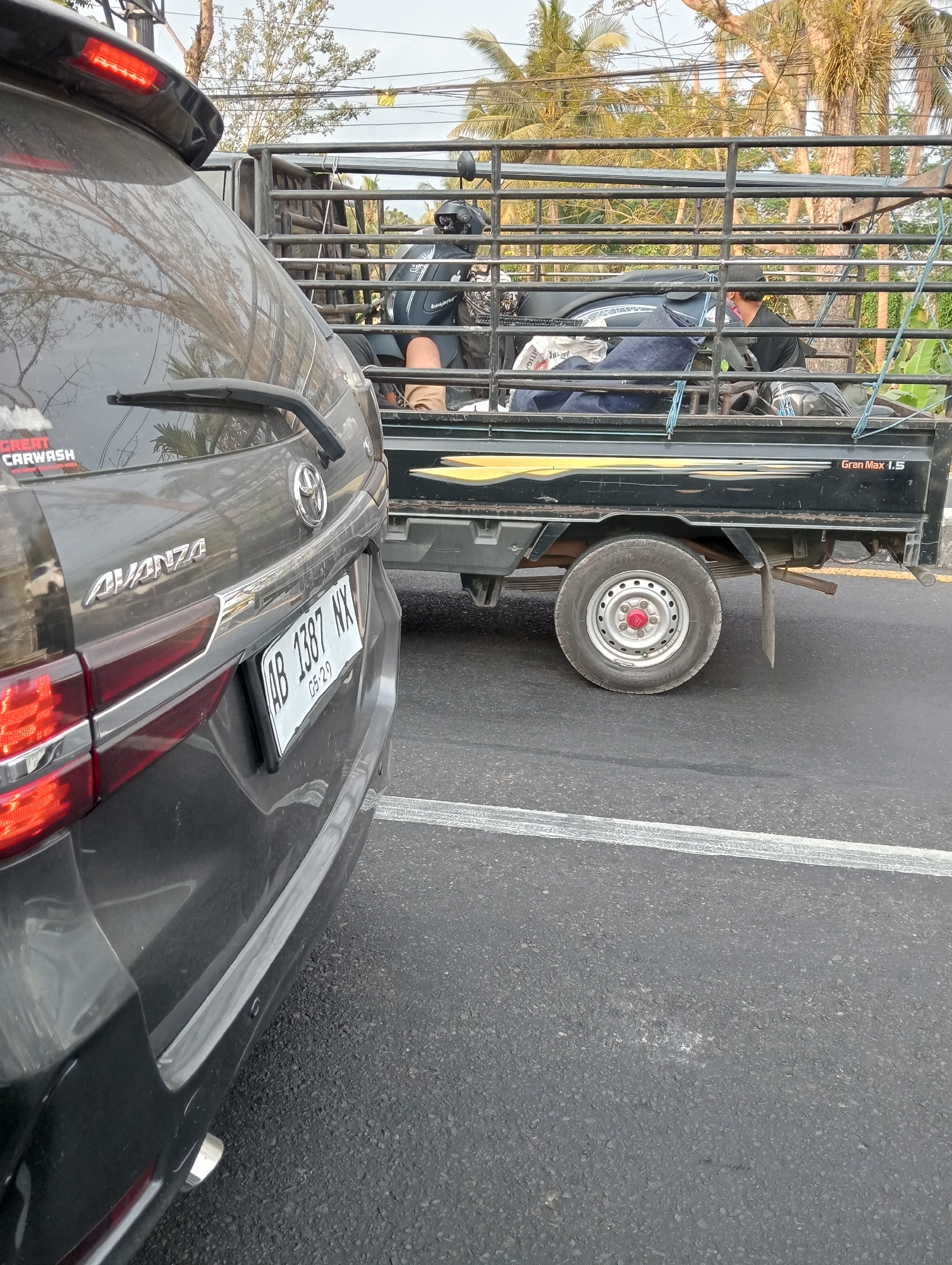 A dark-colored Toyota Avanza is parked on the right side of a two-lane asphalt road. The car's rear is facing the viewer, with the license plate visible: AB 1387 NX, dated 05-29. A black pickup truck, labeled "Gran Max 1.5" on its side, is traveling in the same direction as the Avanza. The pickup truck's bed is covered with a metal cage, and it is transporting a motorcycle and at least one person. The motorcycle, a dark blue scooter, is secured within the cage, and a person wearing a black helmet and dark clothing is seated on it. The background features palm trees and overcast skies, typical of a tropical climate. The overall scene suggests a mundane transportation event on a street in Tempel, Indonesia.
