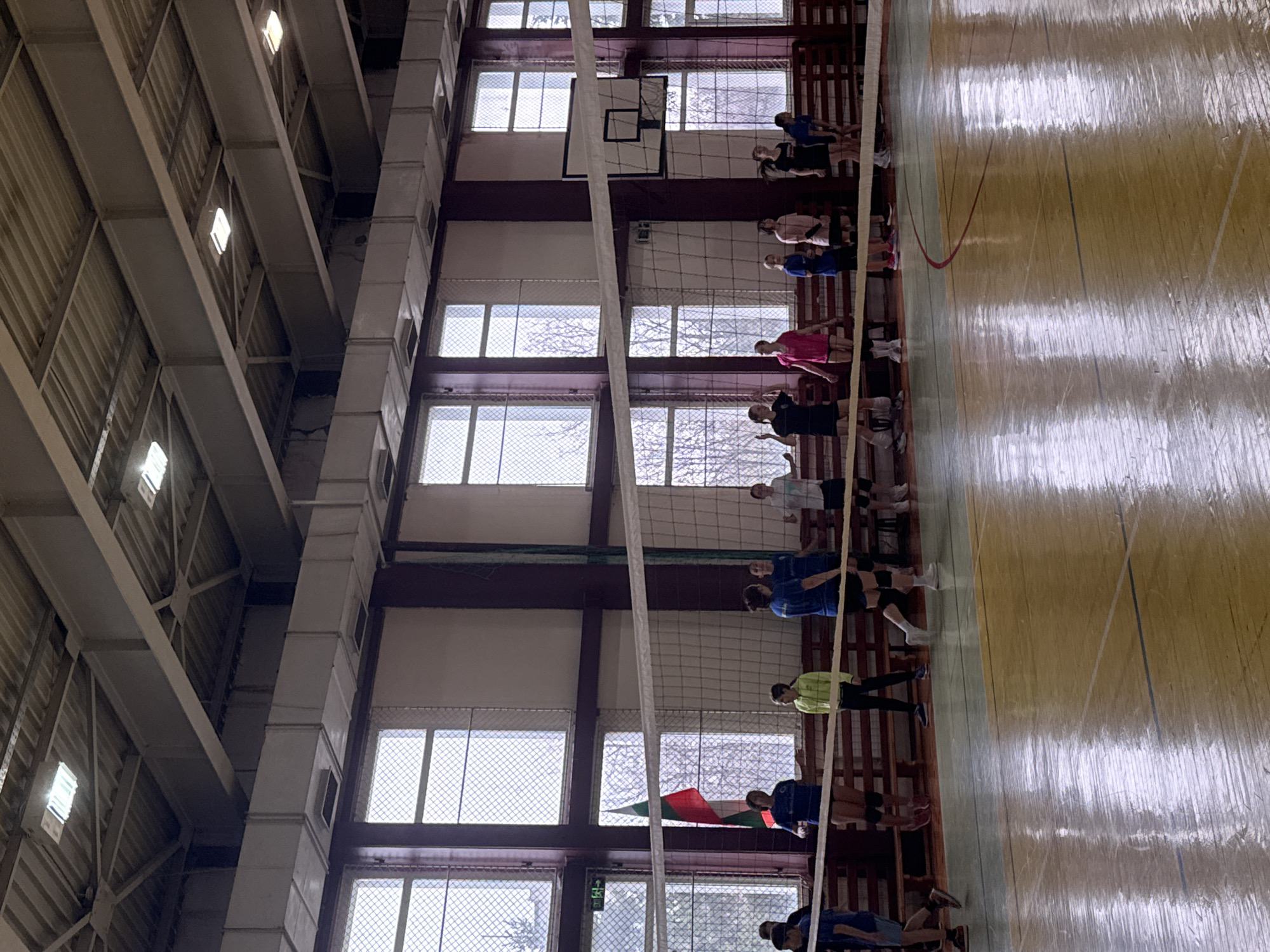 This image depicts an indoor sports gymnasium, located in Sofia, Bulgaria, as evidenced by a Bulgarian national flag visible on the left wall. Approximately 15 to 20 individuals, predominantly young females, are present on and around a volleyball court. Most are dressed in athletic wear, including t-shirts, shorts or leggings, and some wear kneepads.
A white volleyball net spans the width of the court, separating the players into two groups. The court itself features a varnished wooden floor with discernible painted lines in yellow, blue, and red. Several individuals are positioned on both sides of the net, standing or walking, suggesting a pause in play or a practice session.
The gymnasium's far wall is dominated by a series of large, multi-paned windows, each covered with a fine wire mesh or grating. Through these windows, leafless trees and a uniformly overcast sky are visible. Below the windows, wooden benches line the perimeter. An emergency exit sign (green with white pictogram) is mounted near the Bulgarian flag on the left wall. On the right side, behind the volleyball net, a basketball hoop and backboard are attached to a support structure.
The ceiling features exposed metal trusses, corrugated metal panels, several rectangular fluorescent light fixtures, and white rectangular ventilation ducts. The walls are painted in a light color with dark maroon vertical structural beams.