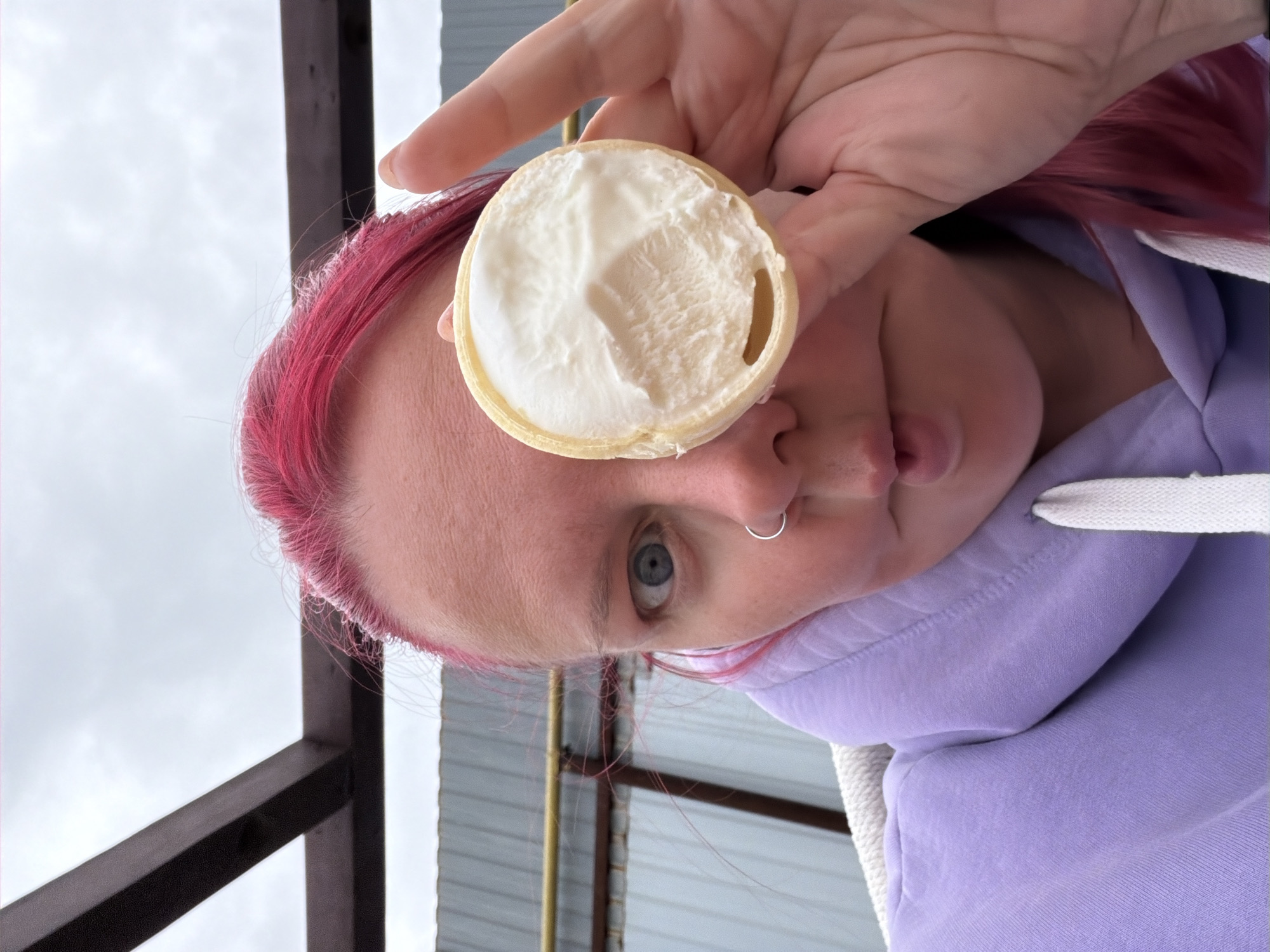An outdoor portrait in the city of Chaikovsky, Russia, depicts an individual with bright magenta hair, pale skin, and a silver hoop piercing in their left nostril. The subject, wearing a lavender hooded sweatshirt with a white drawstring, has their right eye obscured by a light-colored, cylindrical wafer cup. This cup contains a white, textured creamy substance consistent with ice cream, and features a small, circular opening near its base. The individual's left eye, with a visible blue iris, is wide open, and their lips are puckered. The background includes an overcast grey sky, dark brown overhead structural beams, a grey corrugated metal wall, and a horizontal yellow pipe.