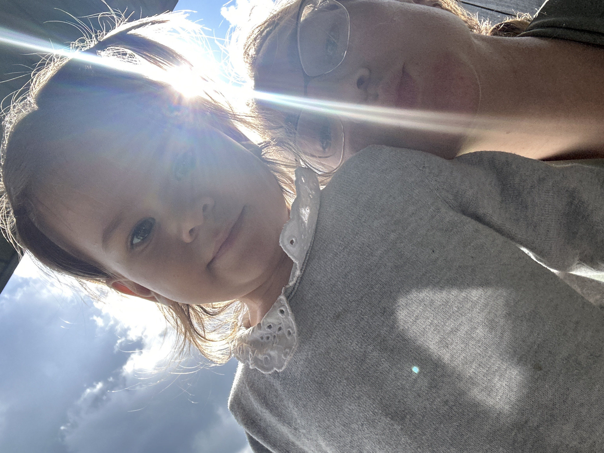 An outdoor selfie features two individuals, a young child and an adult, captured at a low angle against a bright sky in Kharnavy, Russia. The child, a young girl with brown hair, is in the foreground, looking directly at the camera with a neutral expression. She is wearing a grey sweater with a distinctive white, lacy Peter Pan-style collar.
Behind and slightly to the right of the child, an adult, likely a woman, is partially visible. She wears glasses and also looks towards the camera. Both individuals appear to be wearing grey-toned tops.
The image is characterized by strong backlighting from the sun, which is positioned in the upper left, causing prominent lens flare and bright rays of light to stretch across the frame. This intense sunlight creates a halo effect around the subjects' hair and illuminates hair strands, indicating daytime conditions. The background shows a bright blue sky with some white clouds. No specific objects or visible text are present, apart from the subjects and their attire. The overall impression is a casual, sun-drenched moment captured outdoors.