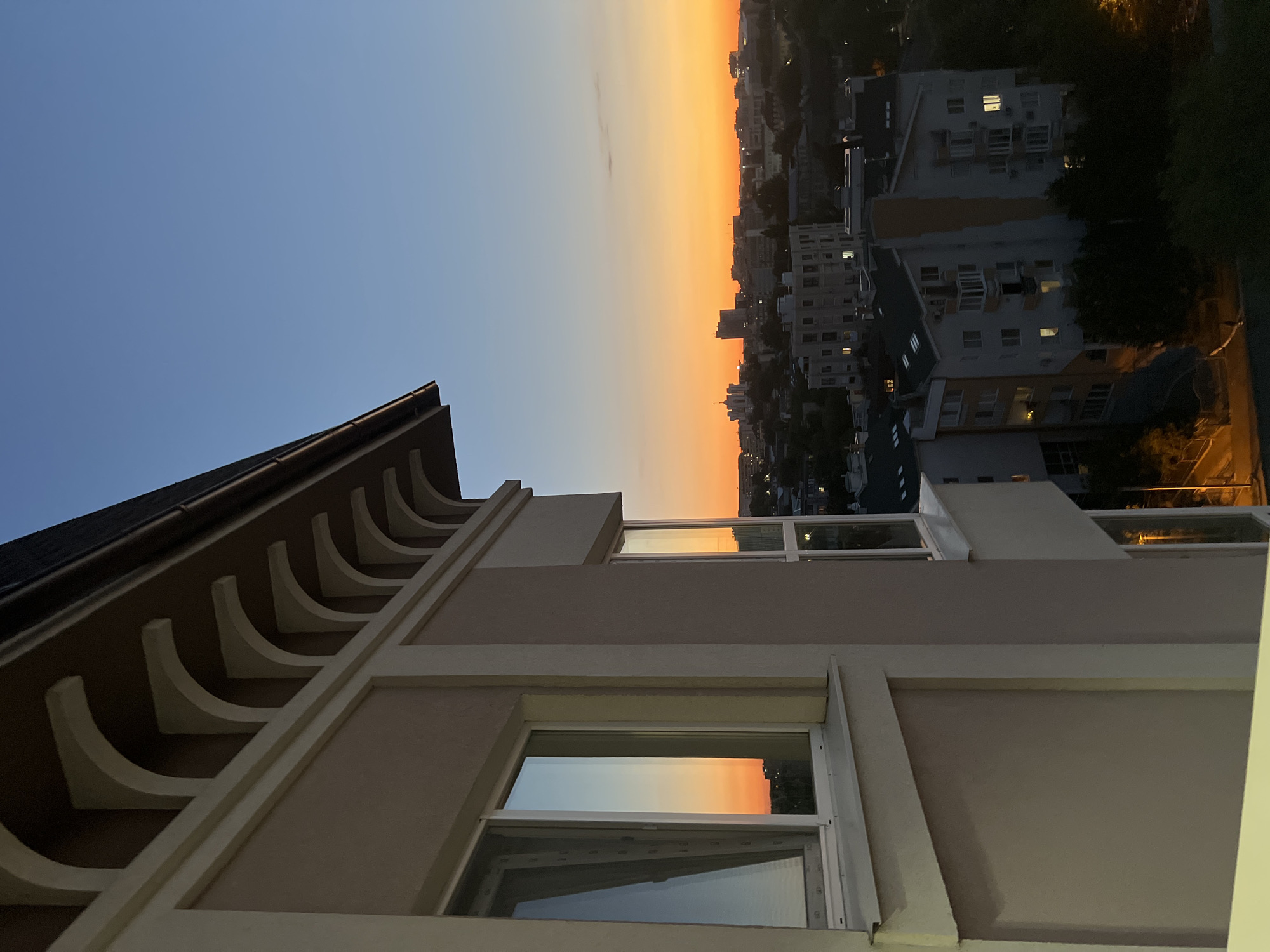 The image captures a twilight view over Chișinău, Moldova, from a residential building. In the foreground, a beige building with decorative arched eaves is visible, featuring several windows reflecting the vibrant orange and blue hues of the sunset. Below, the city sprawls with a mix of apartment buildings, some illuminated with interior lights, and darker, tree-lined areas. The sky transitions from a deep blue overhead to a bright orange band near the horizon, with silhouetted buildings and distant lights creating a city skyline. No people are visible in the image. The scene conveys a calm, urban evening atmosphere.