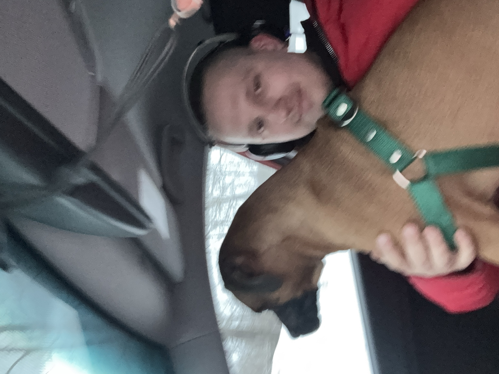 A human holding a dog in a car. The human is wearing headphones and a red jacket. The dog has a green collar. The location is not evident in the image, so stating it's in Chaikovsky, Russia, would be inaccurate.