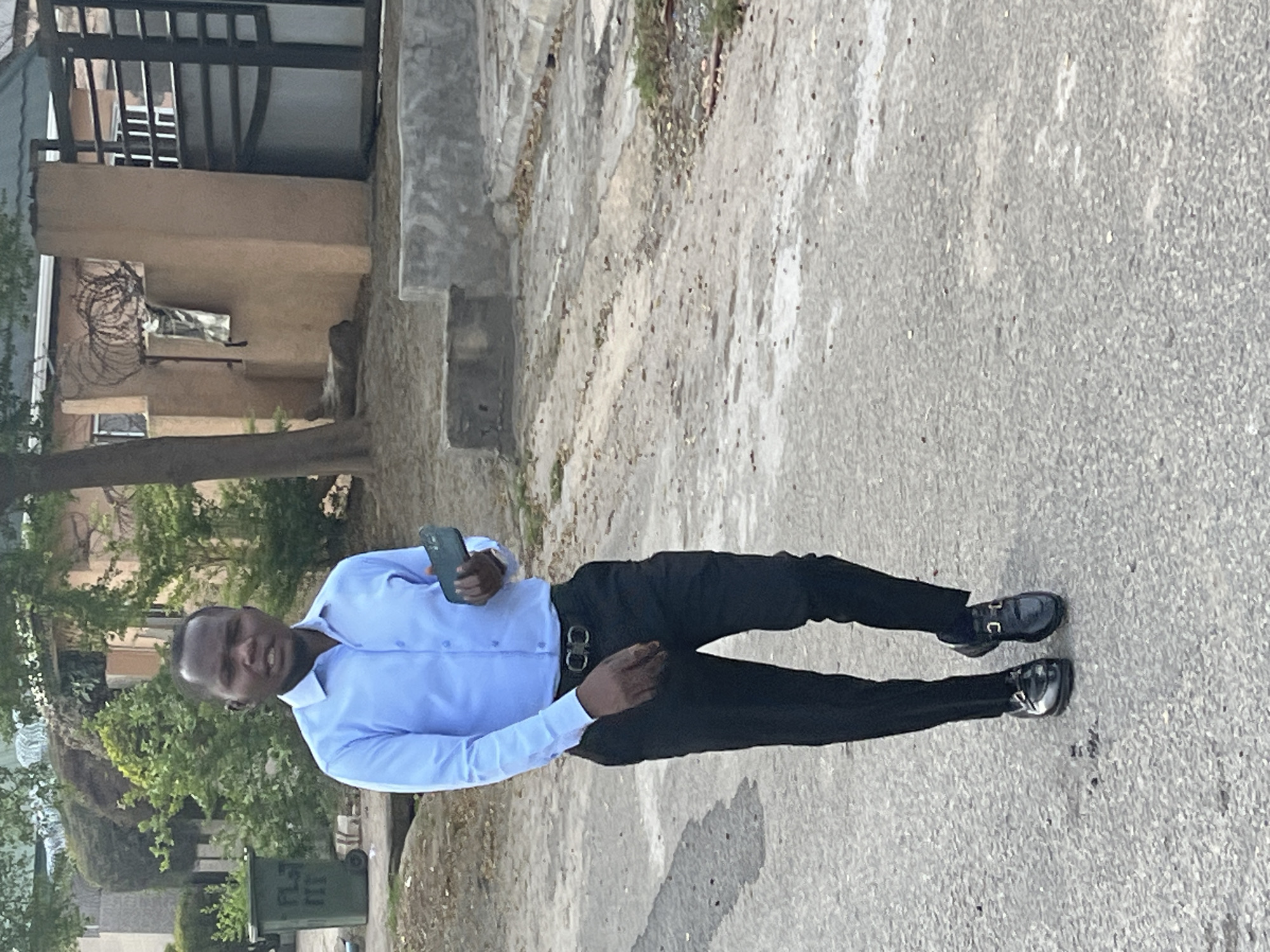 The image captures a young man standing on an asphalt or concrete road during a bright day in Abuja, Nigeria. The man, who appears to be of dark complexion, is positioned centrally, facing slightly right and looking towards the viewer with a neutral expression, his mouth slightly open. He is dressed in a light blue long-sleeved button-up shirt, black trousers, and shiny black dress shoes with silver buckle details. A black belt with a silver-colored buckle is visible around his waist. He holds a dark-colored smartphone in his left hand.
The background features an urban or suburban setting. To the left, behind the man, is a building with a light brown facade and a balcony equipped with dark metal railings. Barbed wire or razor wire is visible along a lower section of the building, possibly for security. Lush green trees and foliage flank the road on both sides. A green, wheeled trash bin with the visible text "PL 11" is partially seen behind the man, near the trees. The road surface is gray and appears slightly textured. The overall lighting suggests clear weather conditions.