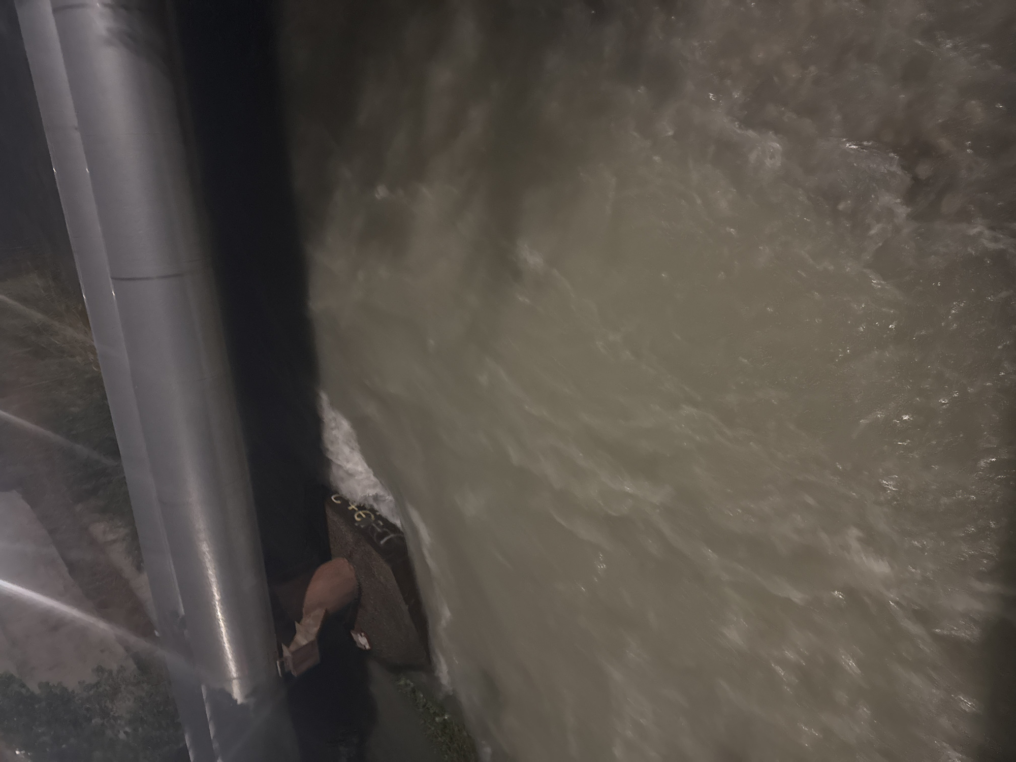 A drone shot, taken from a high angle, shows a section of a large pipe running horizontally across the frame. Below the pipe, a concrete barrier or structure is partially submerged in murky, fast-moving water. The water exhibits turbulence, with white foam and ripples indicating a strong current. On the surface of the concrete structure, there appears to be some text or markings, though they are indistinct due to the water and lighting conditions. The overall environment suggests an industrial or infrastructural area, possibly a drainage channel or canal, within Bishkek City, Kyrgyzstan. The lighting is dim, suggesting it is either dusk, dawn, or nighttime. No individuals are visible in the scene.