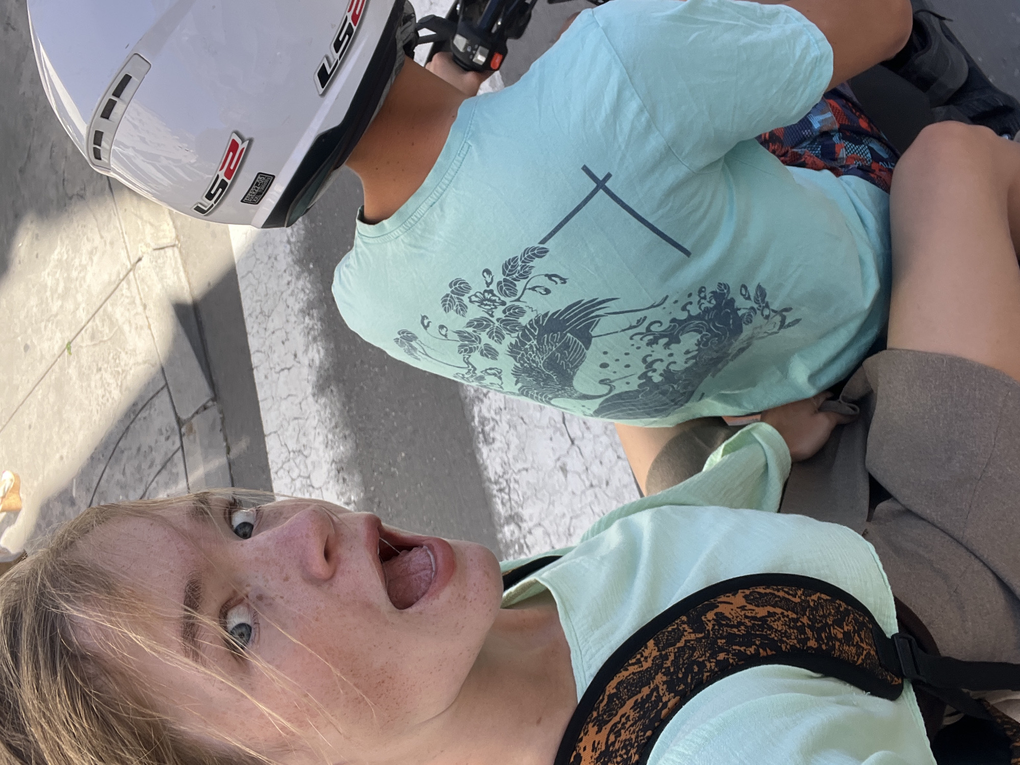 In Budva, Montenegro, two individuals are captured from a high-angle perspective, appearing to be on a motorcycle or scooter. The person in the foreground, a young woman with blonde hair and fair skin, is looking directly at the camera with a surprised or excited expression, her mouth open. She has freckles across her nose and cheeks and is wearing a light green top and a backpack with a distinctive orange and black patterned strap. The second individual, presumably driving, is in the background, facing away from the camera. They are wearing a light blue t-shirt with a dark blue crane and floral graphic on the back, and a white helmet with "LS2" branding. The background shows a paved surface with white markings, suggesting a street or crosswalk. The lighting indicates it is daytime, with shadows cast on the ground.