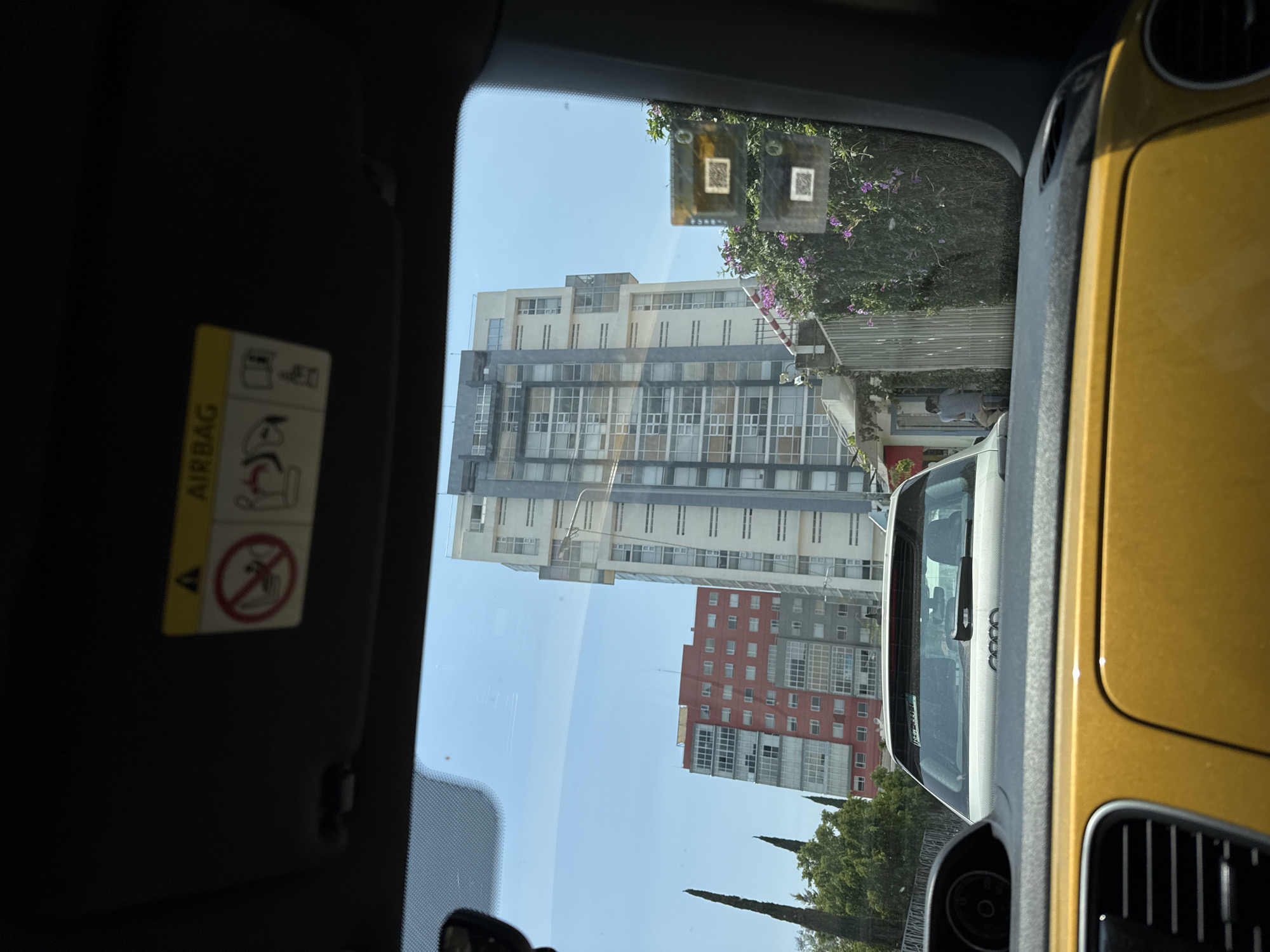 The image, taken from inside a car, shows a view through the windshield towards a cityscape. The location is Puebla, Mexico. Dominating the view are two buildings: one is a tall, modern, multi-story structure with a facade of light-colored panels and numerous windows. The second building, closer to the bottom of the frame, is a red and gray multi-story building with many windows. A yellow car is visible on the right side of the frame, partially blocking the view. A light-colored car is in front of the viewer's car, facing the same direction. In the top portion of the windshield, there are two square stickers with what appear to be QR codes. On the left, a sun visor is down, with an airbag warning label clearly visible. The sky is clear and blue. Green foliage and trees are visible at the bottom.