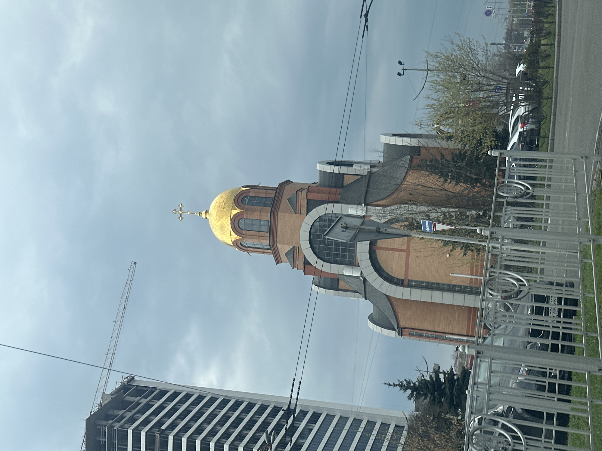 The image depicts a view of a modern-style Orthodox church with a golden dome and cross, situated in Kyiv, Ukraine. The church is constructed of brick and stone, with architectural features including arched windows and decorative elements. Adjacent to the church is a modern, high-rise building under construction, evidenced by a tall construction crane nearby. The scene includes a street with parked cars, a metal fence, and vegetation like trees. The sky is overcast. Wires are strung above.
