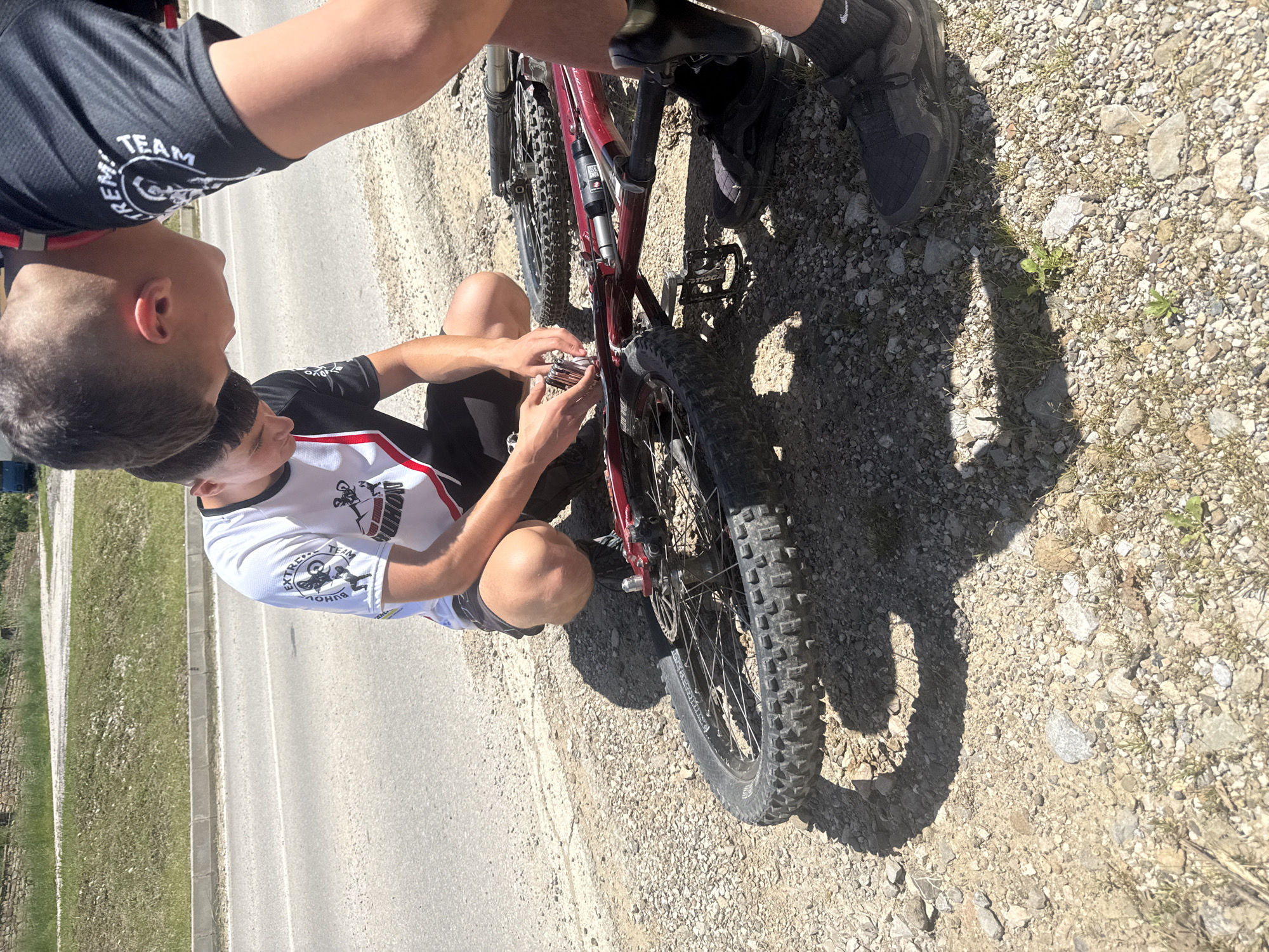Two individuals are repairing a mountain bike on the side of a road. One person is kneeling and using tools near the bike's frame. The other person stands beside the bike, appearing to assist. Both are wearing athletic shirts that are marked with the word 'TEAM' as well as athletic shoes. The mountain bike is red. The location appears to be near Donchovci, Bulgaria, adjacent to a paved road with a gravel shoulder and grassy verge. The surroundings include gravel, small plants, and some vegetation.
