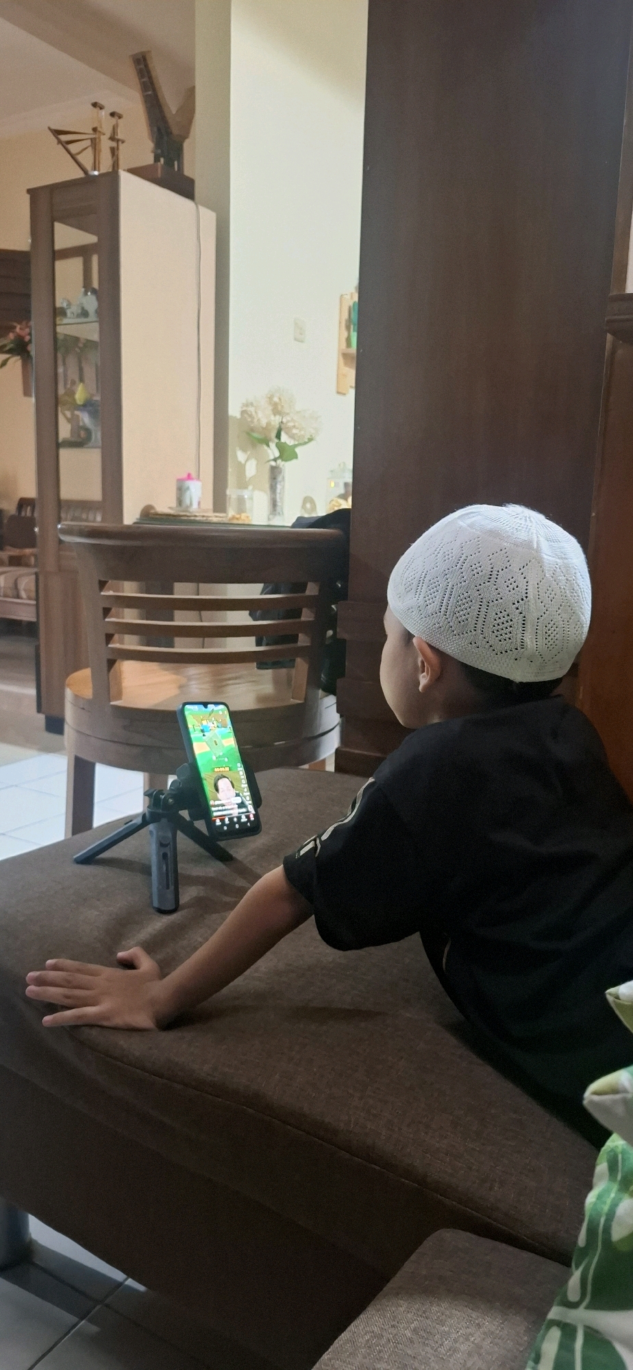 A young boy wearing a white skullcap and a black t-shirt is lying on a brown sofa, looking at a smartphone placed on a small tripod. The phone screen displays what appears to be a video call or streaming content with a person visible on the screen. The boy's right arm is extended, with his hand resting on the sofa cushion. In the background, a wooden cabinet with glass doors is visible, containing decorative items. A wooden chair with a slatted back is positioned in front of the cabinet. The room appears to be a living area in a home, possibly in Desa Gagaksipat, Indonesia, with a light-colored wall and a wooden floor.