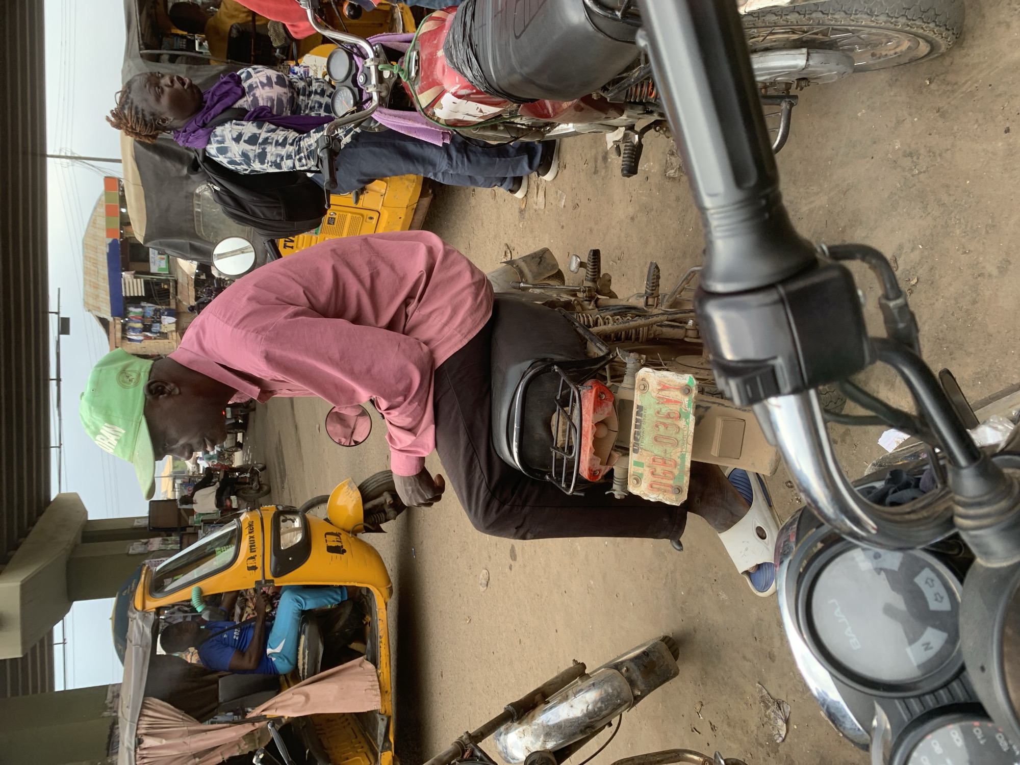 The image captures a street scene in Shagamu, Nigeria, likely at a transportation hub or busy intersection. In the foreground, a man wearing a pink long-sleeved shirt, dark pants, and white and blue slides sits on a black motorcycle. A license plate on the back of his motorcycle reads "OGUN DGB-036YY". To the left, a yellow auto-rickshaw is parked, with a man in a blue shirt and light blue pants visible in the driver's seat. Behind the man on the motorcycle, a woman with braided hair, wearing a patterned top and a purple scarf, stands next to another motorcycle, with her mouth open as if speaking. Her backpack is slung over her shoulders. In the background, other vehicles and buildings are visible, suggesting a bustling urban environment. The ground is unpaved and dusty. Overhead, a concrete overpass is visible, casting a shadow over part of the scene.