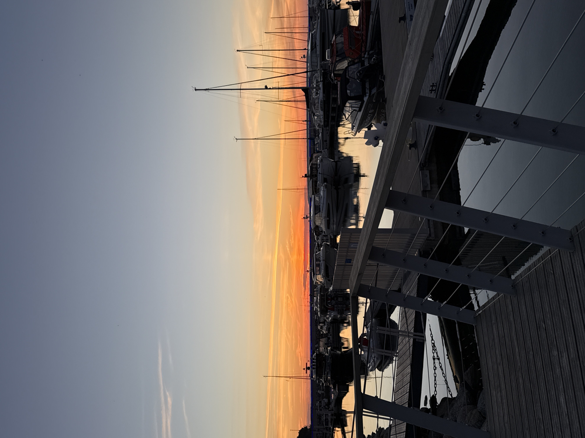 The image depicts a harbor scene at sunset, likely in Tallinn, Estonia, given the subtle architectural cues and the presence of various boats. The sky is a gradient of pale blue at the top, transitioning to vibrant orange and yellow hues near the horizon, indicating the sun has recently set or is setting. Thin, wispy clouds are scattered across the sky, reflecting the warm tones of the sunset.
In the foreground, a wooden walkway with metal railings leads towards the water. The railings are composed of vertical posts and horizontal cables, with a wooden handrail at the top. The walkway itself is constructed of weathered wooden planks.
The water in the harbor is calm and reflects the colors of the sky and the surrounding boats. Several boats are moored in the harbor, ranging from smaller motorboats to larger sailboats with tall masts. The boats are silhouetted against the bright horizon in many cases, but some details are visible, such as cabins and rigging. One boat in the mid-ground on the right appears to be a red and white rescue vessel. Another smaller dinghy is visible closer to the foreground, tethered to the walkway.
In the background, a faint line of trees or land is visible along the horizon, further accentuated by the sunset. The overall impression is one of tranquility and natural beauty as day transitions into evening. No people are visible in the frame. The environment is a marina, suggesting a recreational or commercial boating area.