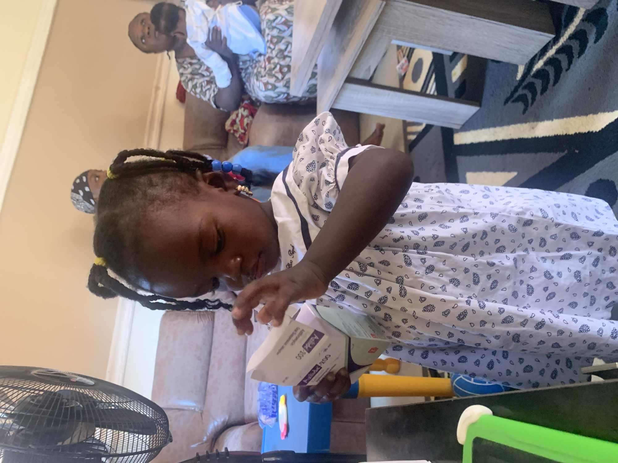 The image depicts an indoor scene with several people and objects. In the foreground, a young Black girl with braided hair, adorned with blue beads and yellow accents, is leaning forward and holding a small white and purple box. She is wearing a white dress with a dark blue paisley pattern. To her left, a portion of a black oscillating fan is visible.
In the background, to the left of the girl, an adult Black woman in a patterned dress is holding a baby dressed in white. They are seated on a couch. Behind them, a wooden table with a dark, patterned rug is visible. The room appears to be a living area, suggesting a casual, domestic setting. The lighting suggests it is daytime.
The activity appears to be a family gathering or a moment of interaction between the individuals, with the young girl examining the contents of the box. The overall mood seems relaxed and familial.
The visible text on the box the girl is holding reads "Axiltea 200" in black lettering.