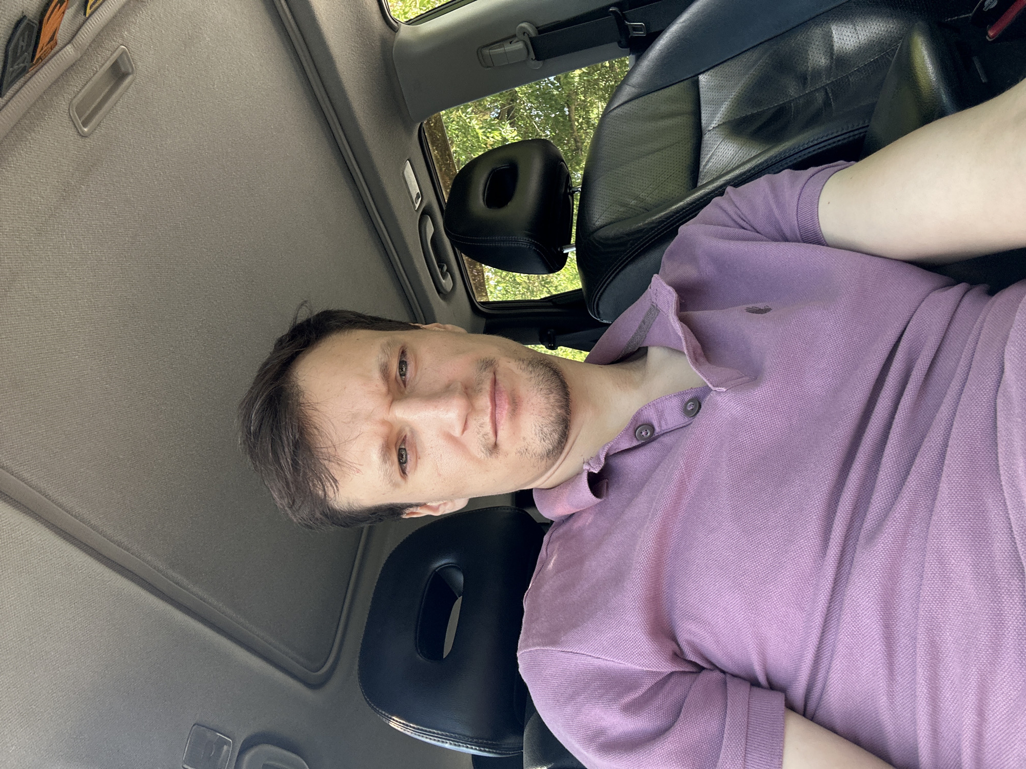 A man is seated inside a car, likely a Nissan X-Trail, judging by the headrest design. He is looking directly at the camera, with a slightly serious expression. The car's interior features black leather seats and a grey fabric headliner with a sunroof. Outside the car, lush green trees are visible through the window, suggesting a daytime setting with natural light. The man is wearing a lavender polo shirt with two buttons and a small embroidered logo on the left side of his chest. The image is a selfie, taken from a slightly low angle, which makes the car's ceiling and sunroof prominent. The specific location is identified as Makiv, Ukraine, based on the provided context, although no explicit visual cues within the image confirm this.