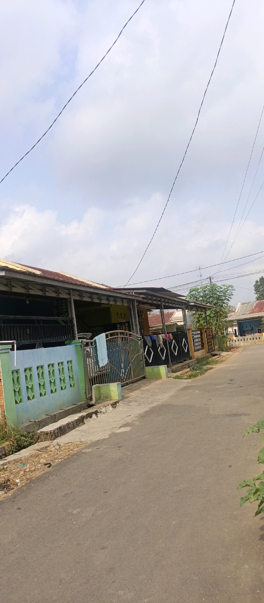 The image shows a residential street scene in Kotabumi, Indonesia, under a cloudy sky. A paved road runs from the foreground into the distance, bordered by a concrete drainage channel and vegetation on the left, and a fence and houses on the right. The houses appear to be single-story structures with corrugated metal roofs supported by visible beams. In the foreground, a light blue fence with decorative cutouts runs along the property line, adjacent to a metal gate. A light blue towel is hanging on the gate. Beyond the gate, clothes are drying on a line in front of another section of fence. The environment suggests a typical neighborhood with modest housing and utility lines overhead.