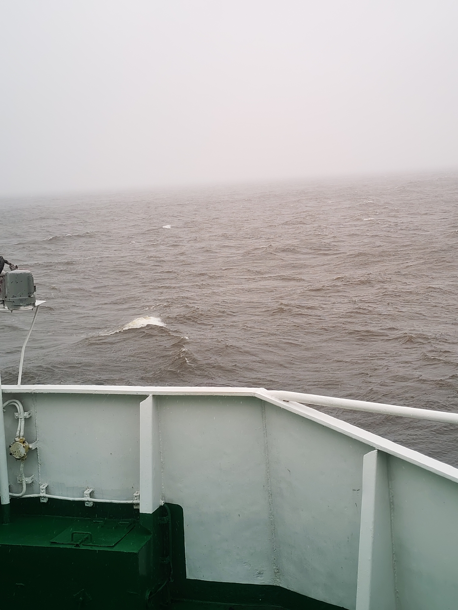 The image presents a view from the deck of a ship traversing a large body of water under dense fog or mist. The scene is characterized by muted grays and whites, indicating low visibility and an overcast atmosphere.
In the foreground, the sturdy, light-colored metal structure of the ship's deck and railing is prominent, with a section of the deck painted dark green. Visible on the left side of the deck is some machinery or equipment, possibly part of a winch or mooring system, connected by white pipes or conduits. Small water droplets are discernible on the ship's surfaces, suggesting rain or heavy moisture in the air.
Beyond the ship, the expansive water appears choppy and dark, with numerous waves and whitecaps breaking across the surface, particularly in the mid-distance. The horizon line is completely obscured by the thick, featureless mist that merges with the sky, giving the impression of an infinite, hazy expanse. No other ships or landmasses are visible, emphasizing the feeling of isolation.
The weather conditions appear challenging, with poor visibility and rough seas. The lighting suggests daytime, though the heavy overcast makes it difficult to ascertain a specific time. The setting is likely a large lake or coastal area near Shcheleiki, Russia. There are no people visible in the image, and no discernible text.