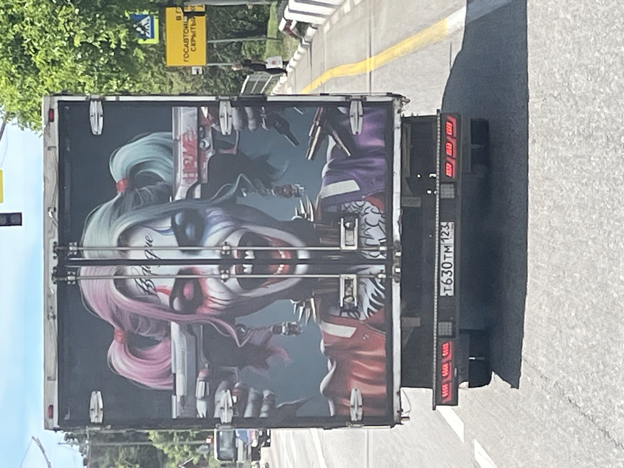 The image shows the rear of a white truck with a large graphic on its doors. The graphic depicts a stylized, zombie-like character with pink and blue pigtails, white face paint with red and blue accents, and a wide, open mouth. The character is holding two pistols, one in each hand, pointed outwards. Text appears on the character's forehead and on the truck doors. The truck is on a paved road, likely a highway, with a yellow line marking the lanes. In the background, there are trees and a signpost with Cyrillic lettering. A pedestrian can be seen on the right side of the road, carrying a white bag. The license plate of the truck is visible, reading "T630TM 123". This scene is located in Dagomys, Russia.