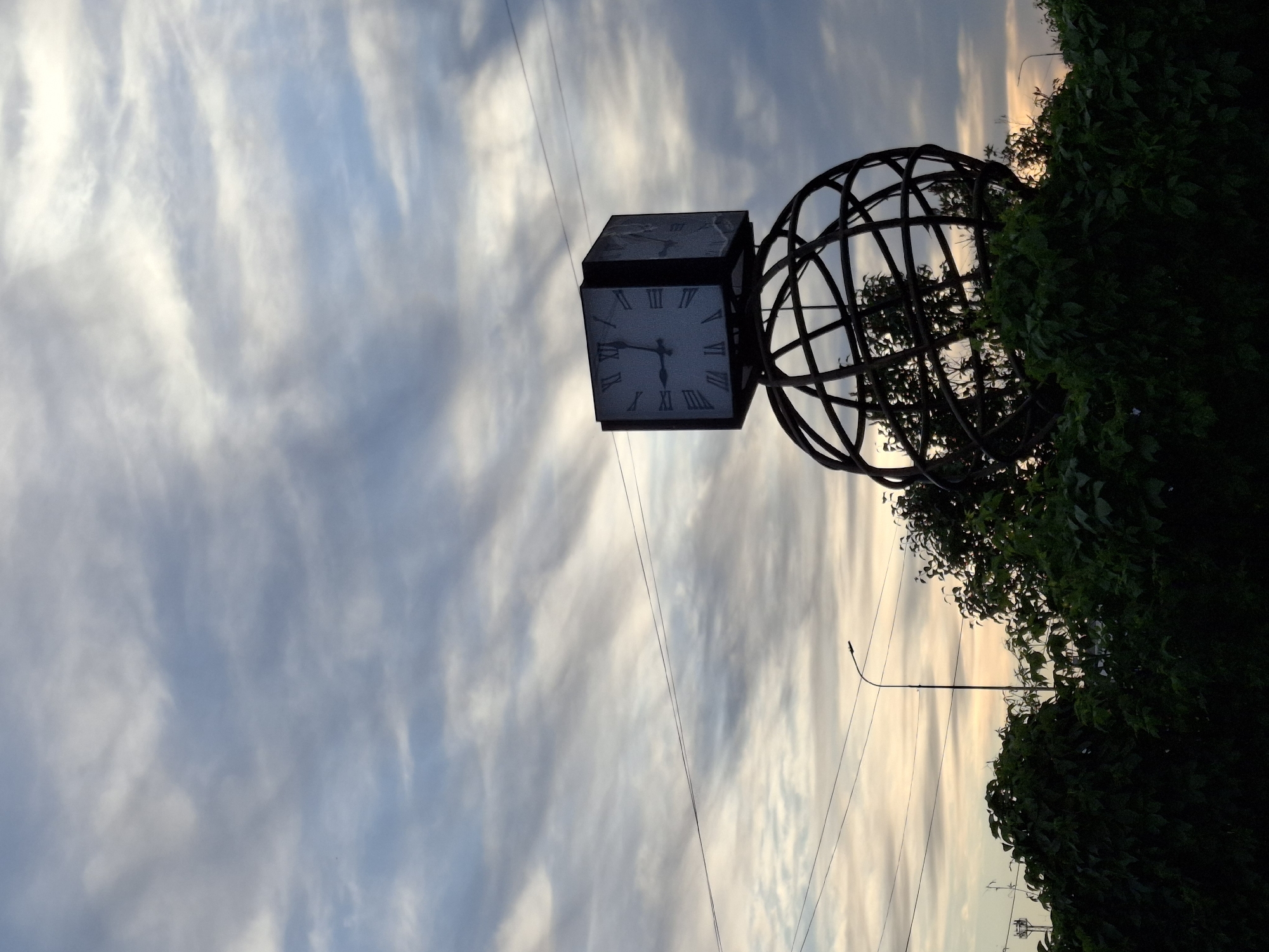 A low-angle, slightly rotated view captures a public art installation in Chaikovsky, Russia. The installation consists of a large, open-framework globe attached to a four-sided clock in a metal frame. The clock faces feature Roman numerals. The time shown is 8:57. The installation is partially obscured by dense green foliage at the right. Overhead power lines traverse the scene against a backdrop of a dynamic sky filled with streaks of blue, gray, and white clouds. A tall, slender lamppost is visible on the right side of the frame near the horizon.