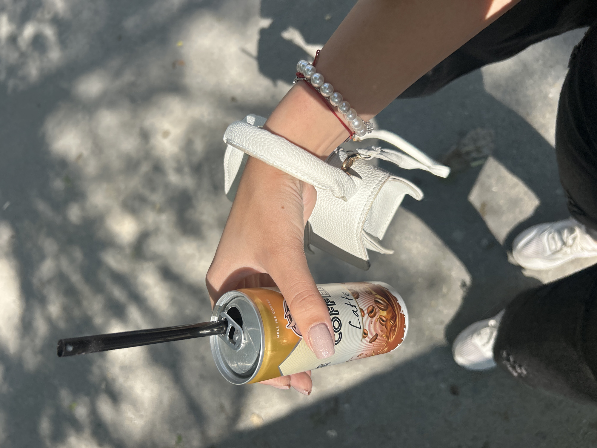 A person is holding a can of "Coffee Latte" with a black straw in it. The person has a white purse resting on their arm and is wearing a pearl bracelet with a red thread. The person is wearing black pants and white shoes. The background is a grey concrete surface with shadows of trees. The location is Unknown, Unknown.
