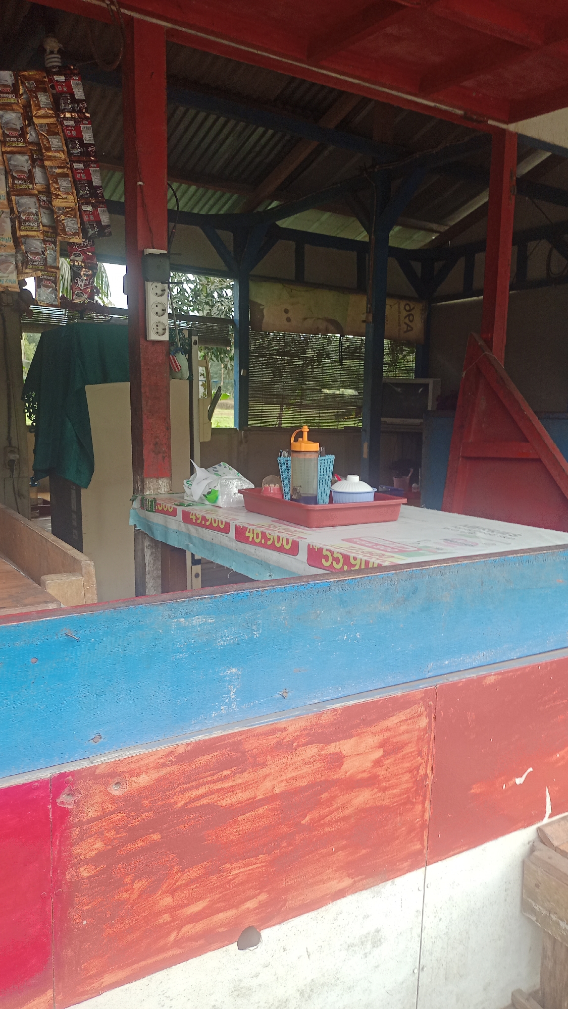 The image depicts an outdoor food stall located in Cikalong, Indonesia. The stall has a red and blue painted wooden counter in the foreground, with a table set up behind it. On the table are several items, including a red tray, a blue plastic container with cutlery, a bottle with an orange cap, a white lidded bowl, and a white plastic bag. Behind the table, a shelf holds packets of what appear to be coffee or instant noodles. The stall is covered by a corrugated metal roof supported by red and blue painted posts. In the background, there are windows with bamboo blinds and some vegetation visible. The overall scene suggests a simple, functional food service establishment in a semi-rural or local market setting. There are no people visible in the image.