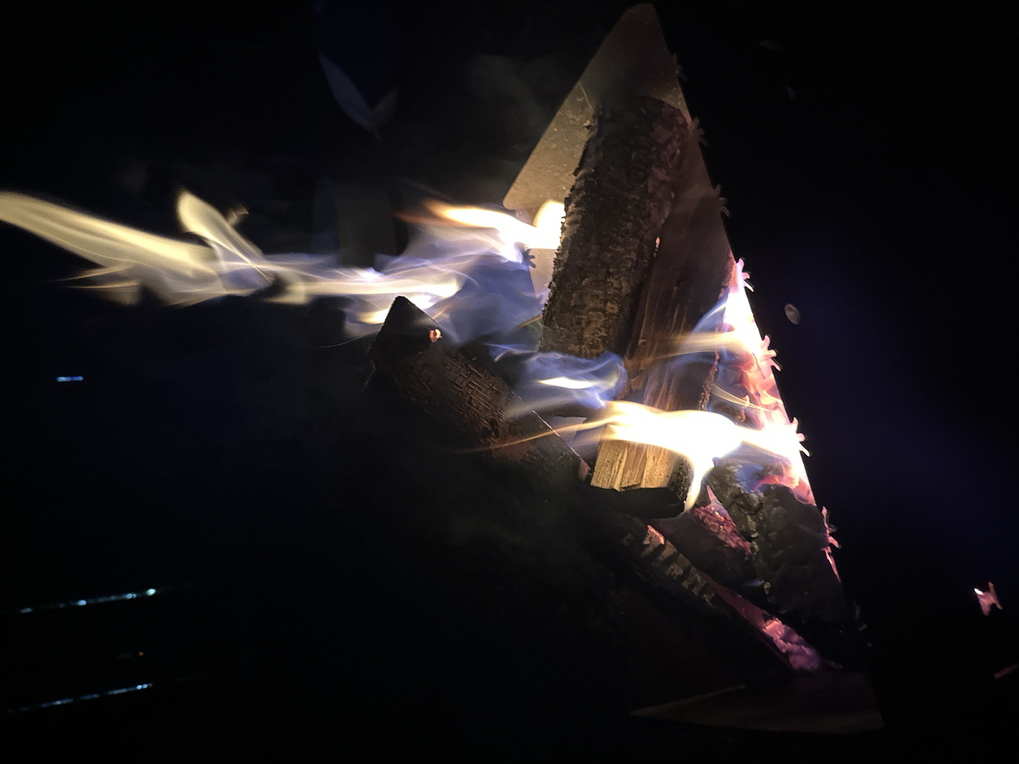 A nighttime scene depicts a campfire. Several logs are stacked, arranged to promote burning. Flames are visible, exhibiting various colors from yellow to blue, indicating the heat of the fire and combustion of different materials. The surroundings are dark, suggesting an outdoor setting at night. There are reflections in the bottom left, possibly from a nearby surface. The exact location in Biysk, Russia, cannot be determined from the image alone.