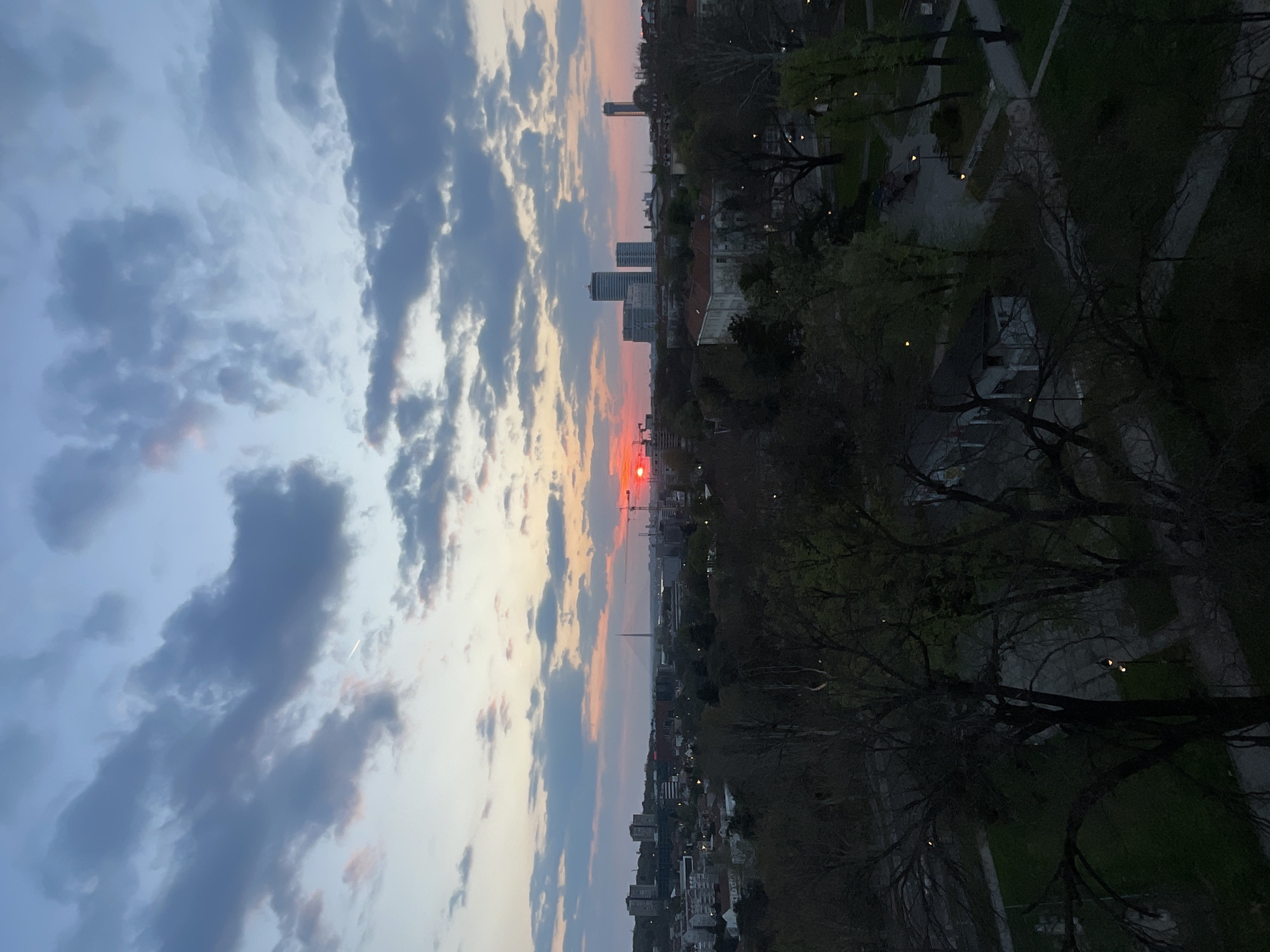 The image shows a sunset over Belgrade, Serbia. In the center of the frame, a red sun is setting behind buildings. To the left, the sky is filled with blue and gray clouds. To the right, there is a view of trees and buildings. In the background, two tall buildings can be seen in the distance, along with the Belgrade Tower.