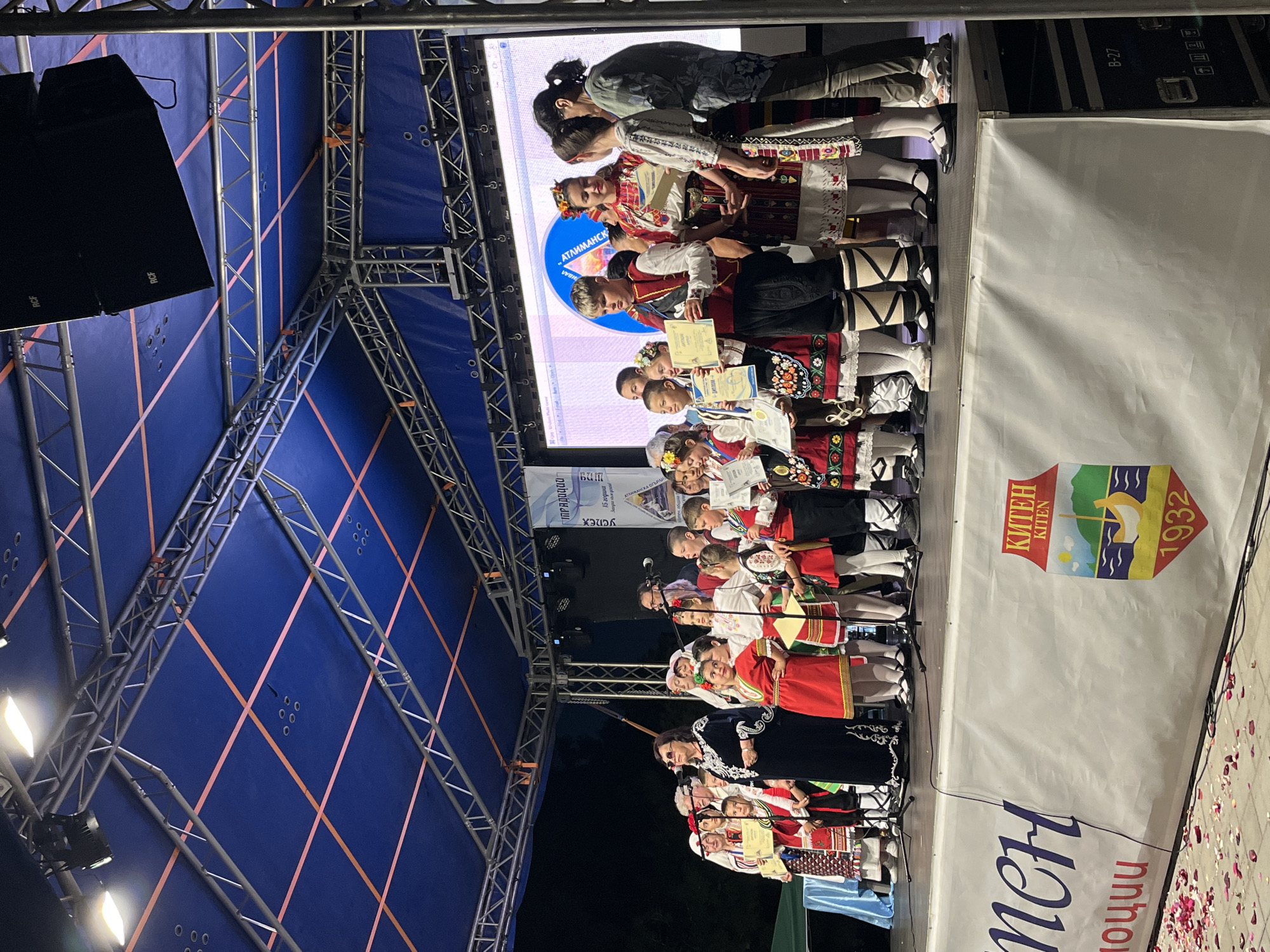 The image captures a group of children and adults on a stage, likely during an awards ceremony or cultural event, in Kiten, Bulgaria. The stage is set against a dark background, with a blue tarp overhead supported by a metal truss structure. Several spotlights are visible on the truss.
In the foreground, a white banner with text and a logo stretches across the front of the stage. The logo is a shield shape with a sailboat, a body of water, mountains, and the text "КИТЕН" (Kiten) and "1932". The text on the left side of the banner is partially visible, reading "мен" and "оции". Scattered on the ground in front of the banner are rose petals.
On stage, a diverse group of individuals are present. Many of the children are wearing traditional Bulgarian folk costumes, characterized by vibrant colors, embroidered patterns, and distinct headwear. Some of the children are holding certificates or awards. An adult woman in the center, wearing a dark outfit with white embroidery, appears to be addressing the audience or participants. Other adults are standing on the stage, some holding microphones.
The lighting on stage is a mix of artificial lights and the ambient evening light, creating a brightly lit performance area. The overall impression is one of a festive community event celebrating cultural heritage.