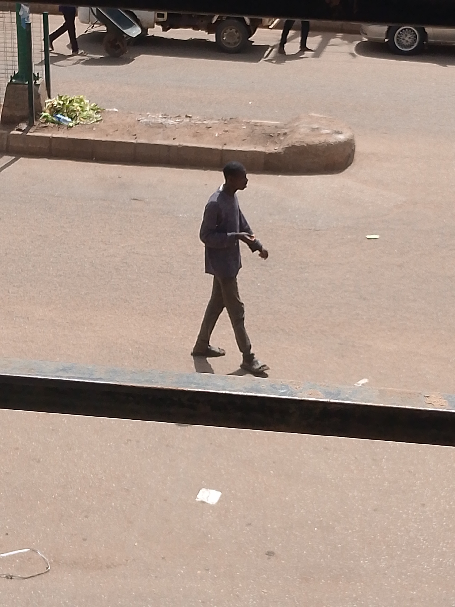 A young man walks across a paved road in Bukuru, Nigeria. He is wearing a dark blue shirt and dark pants, and is looking down. He appears to be walking alone. In the background, a white truck is parked, and there is a pile of green foliage on the side of the road. The sun is shining, indicating it is daytime. The scene appears to be a public street, possibly in a market area, given the presence of the truck and the foliage. The man's posture suggests he is focused on his path, and no particular emotion is discernible. There is no visible text in the image.