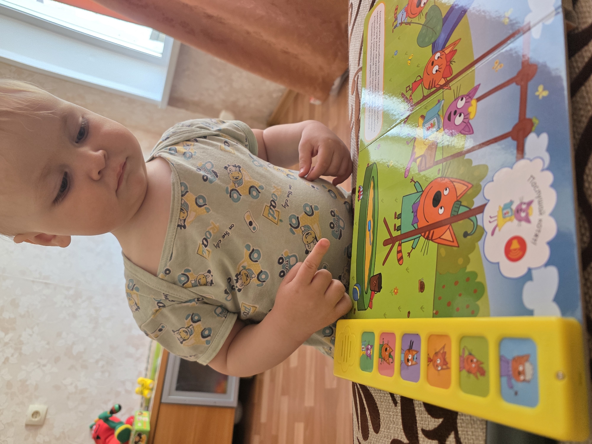 A toddler wearing a light green onesie with vehicle patterns is standing next to a large, colorful children's book. The book has cartoon cat characters and a yellow electronic sound module with push buttons featuring the same characters. The child is pointing at the yellow module. The background includes a light-colored wall, a window, and a wood-patterned floor. The location appears to be an indoor play area.