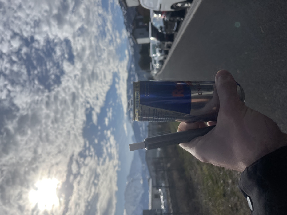 The image presents a close-up perspective of a light-skinned individual's right hand, clad in a dark sleeve, holding two distinct objects in Sallanches, France. The hand firmly grips an opened silver and blue Red Bull energy drink can, featuring French text indicating "BOISSON ENERGISANTE" and "SUCRÉE & TRES FRAIS." Positioned to the left of the can, between the thumb and forefinger, is a slender, metallic grey object with a white tip, consistent with a one-hitter pipe.
The background reveals a sunny outdoor environment under a partly cloudy sky, with a bright sun positioned in the upper left quadrant. Prominently in the distance are snow-capped mountains. Below the mountains, a blend of industrial-type buildings on the left and a residential structure with a dark roof on the right are discernible. A paved path or road extends into the mid-ground on the right, alongside which a linear arrangement of parked vehicles, including cars and vans, recedes into the distance. The immediate foreground on the left consists of dry ground with sparse vegetation.