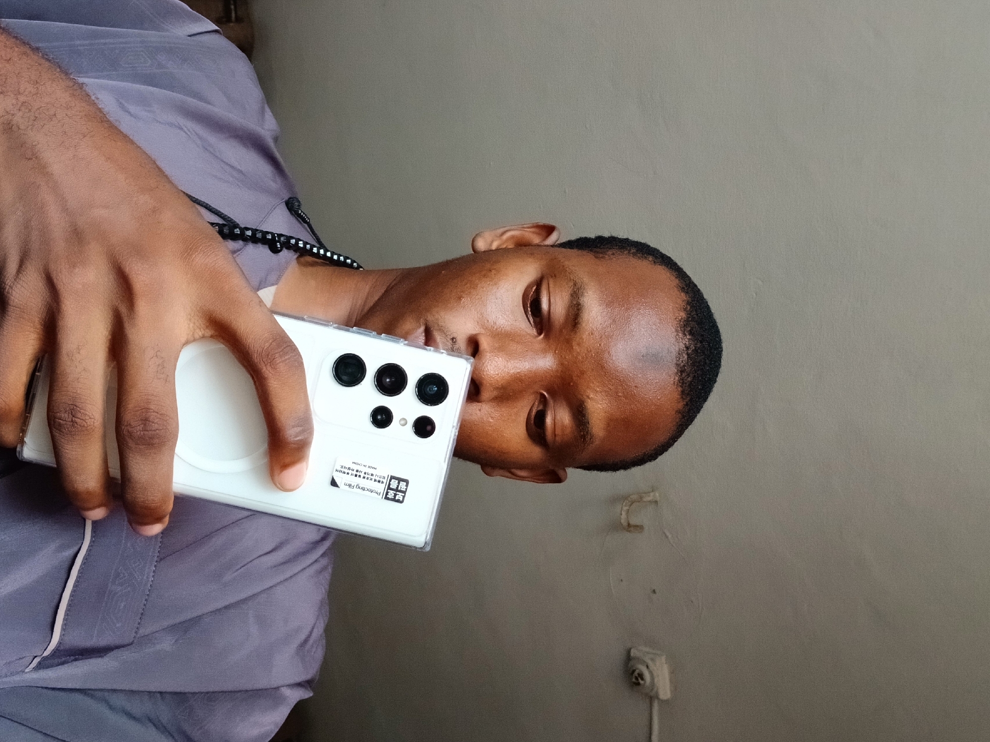 A person holds a white smartphone in a clear case with multiple camera lenses. The phone has a sticker on the back. The person is wearing a purple shirt and a beaded necklace. The background is a plain wall with an electrical outlet. The location is Lagos, Nigeria.