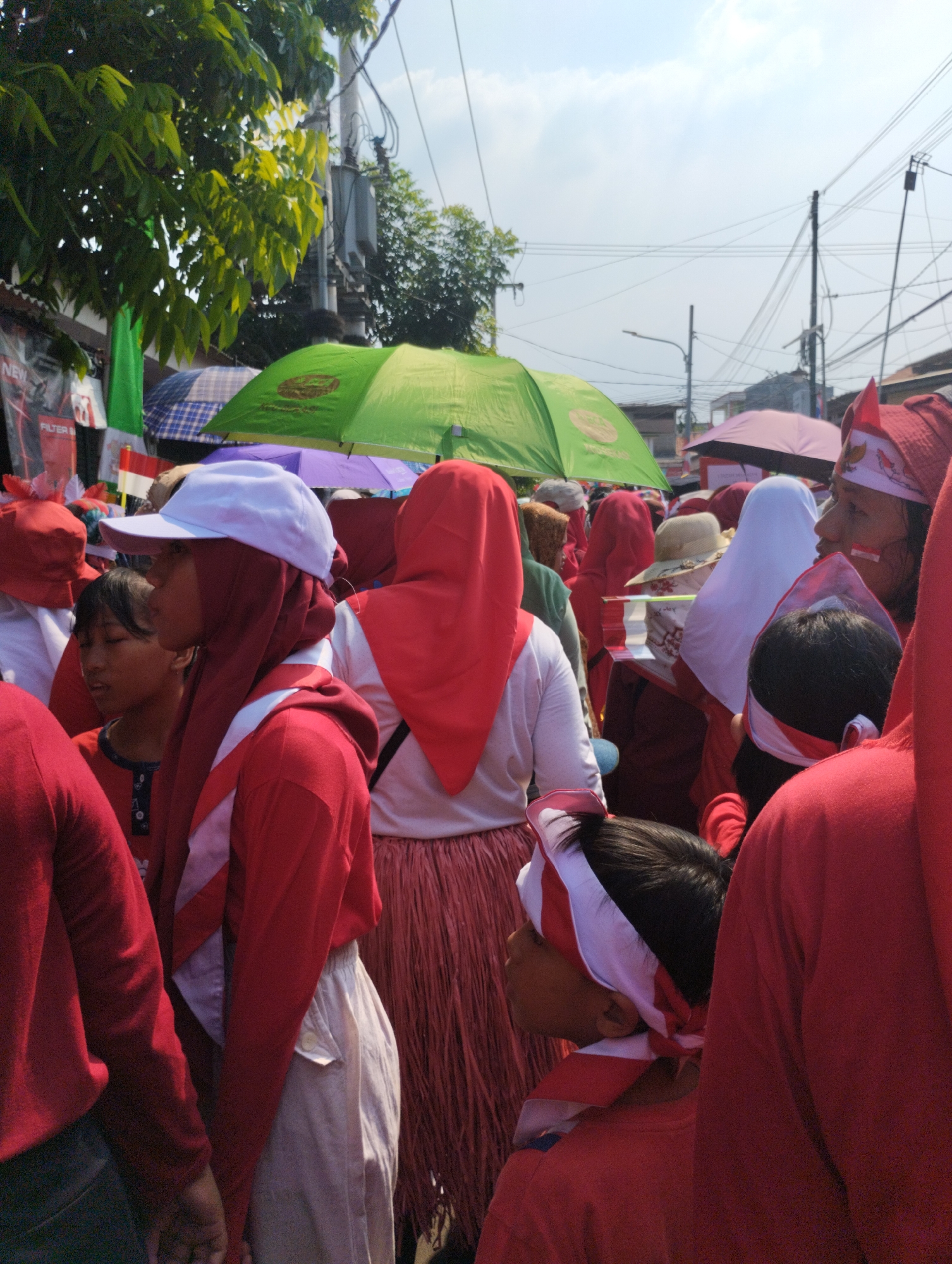 The image captures a crowd of people, predominantly wearing red and white attire, on a street in Bekasi, Indonesia. The atmosphere appears festive, with several individuals sporting headscarves or bands in red and white, possibly indicating a celebration or event. Umbrellas, both green and pink, are visible, suggesting protection from the sun. Various flags, including the Indonesian flag, are also present. The background reveals urban elements such as utility poles, wires, and buildings, characteristic of a city street. The focus is on the group of people, their clothing, and the festive elements, with the urban environment providing the context for the scene.