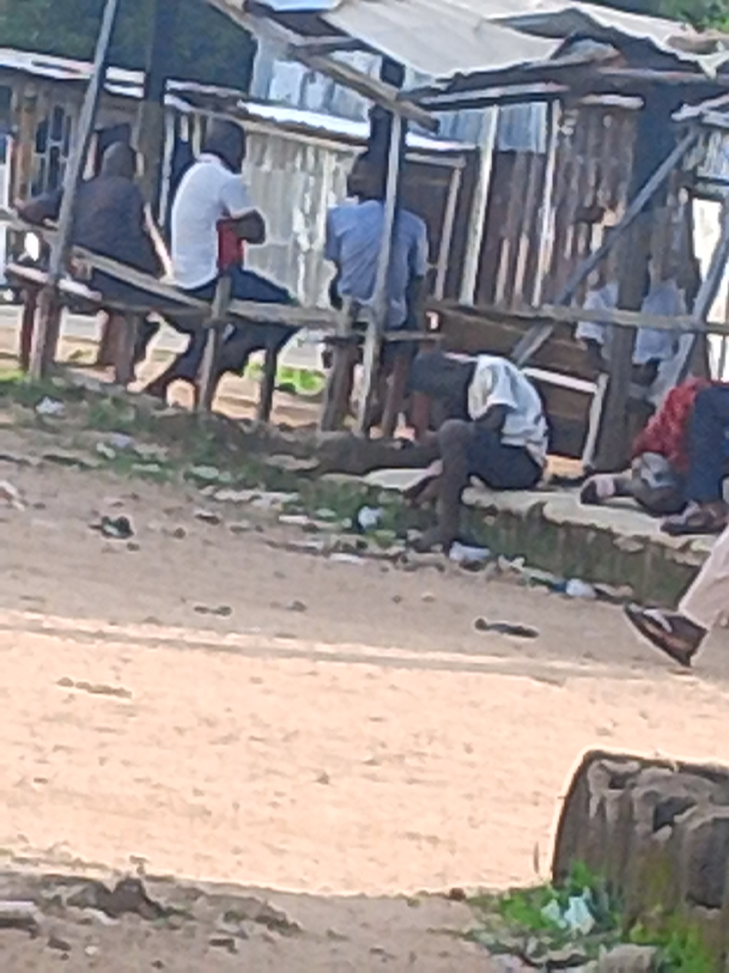 The image depicts an outdoor scene with several individuals gathered under a makeshift shelter. The shelter appears to be constructed from corrugated metal and wood, suggesting a rural or informal setting. The ground is dusty and uneven, with some debris scattered around.
There are at least five people visible in the image. Some are seated on benches or a raised platform under the shelter, while others are positioned on the ground. One person, wearing a white shirt and dark pants, is sitting hunched over, possibly engaged in an activity not clearly discernible. Another person in a light blue shirt is also seated. The lighting suggests it is daytime, possibly late afternoon, given the shadows.
The overall atmosphere seems relaxed, with people resting or socializing in the shade. There are no obvious signs of urgency or significant events taking place. The location, identified as Jalingo, Nigeria, provides a geographic context. There is no visible text in the image. The scene appears to be a communal gathering space, possibly a market stall or a rest area.